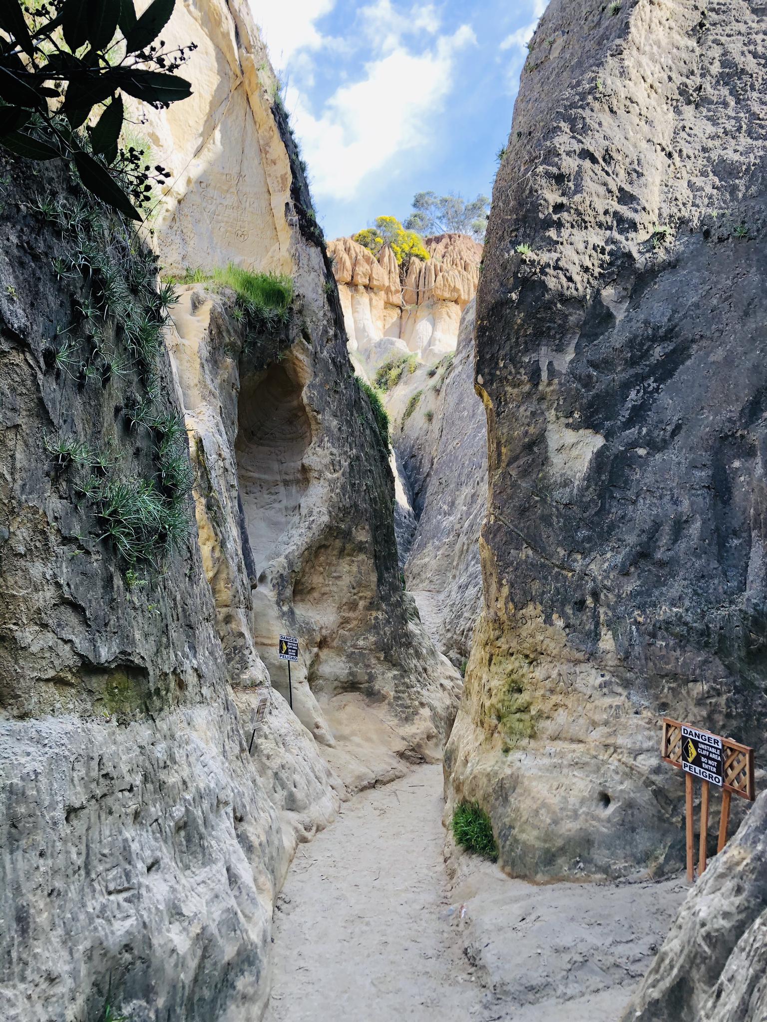 Narrow slot canyon with sandy path and tall rock walls, bright sky.