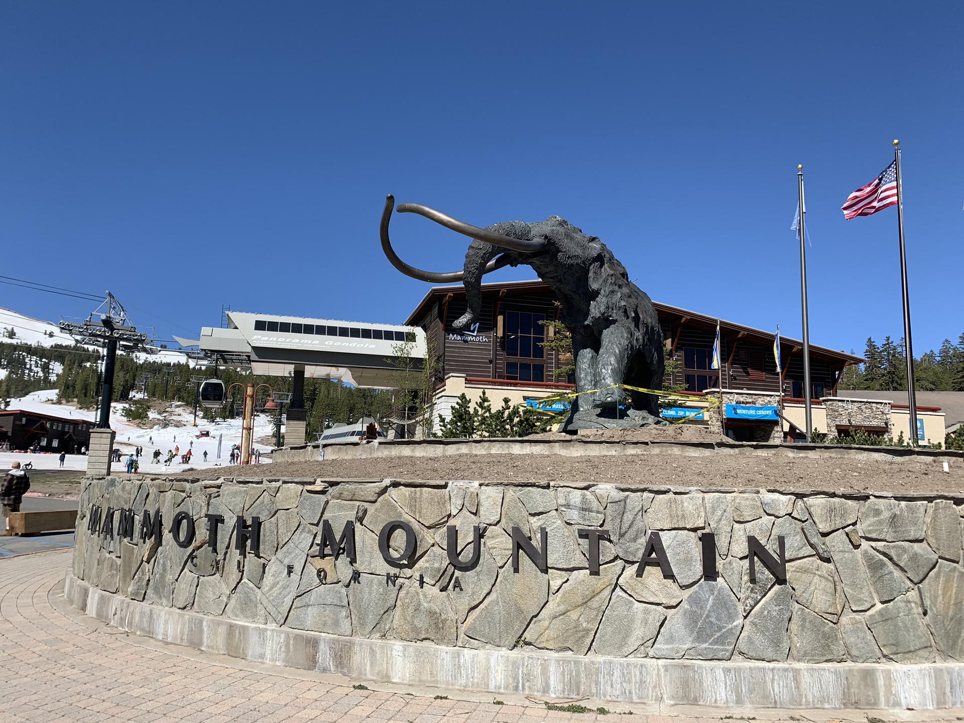 Large bronze mammoth statue on a stone base with buildings and snowy mountains.
