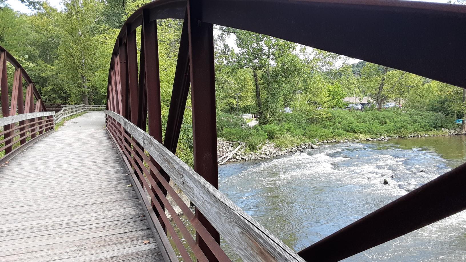 A wooden and metal bridge curves over a flowing river on a bright day.
