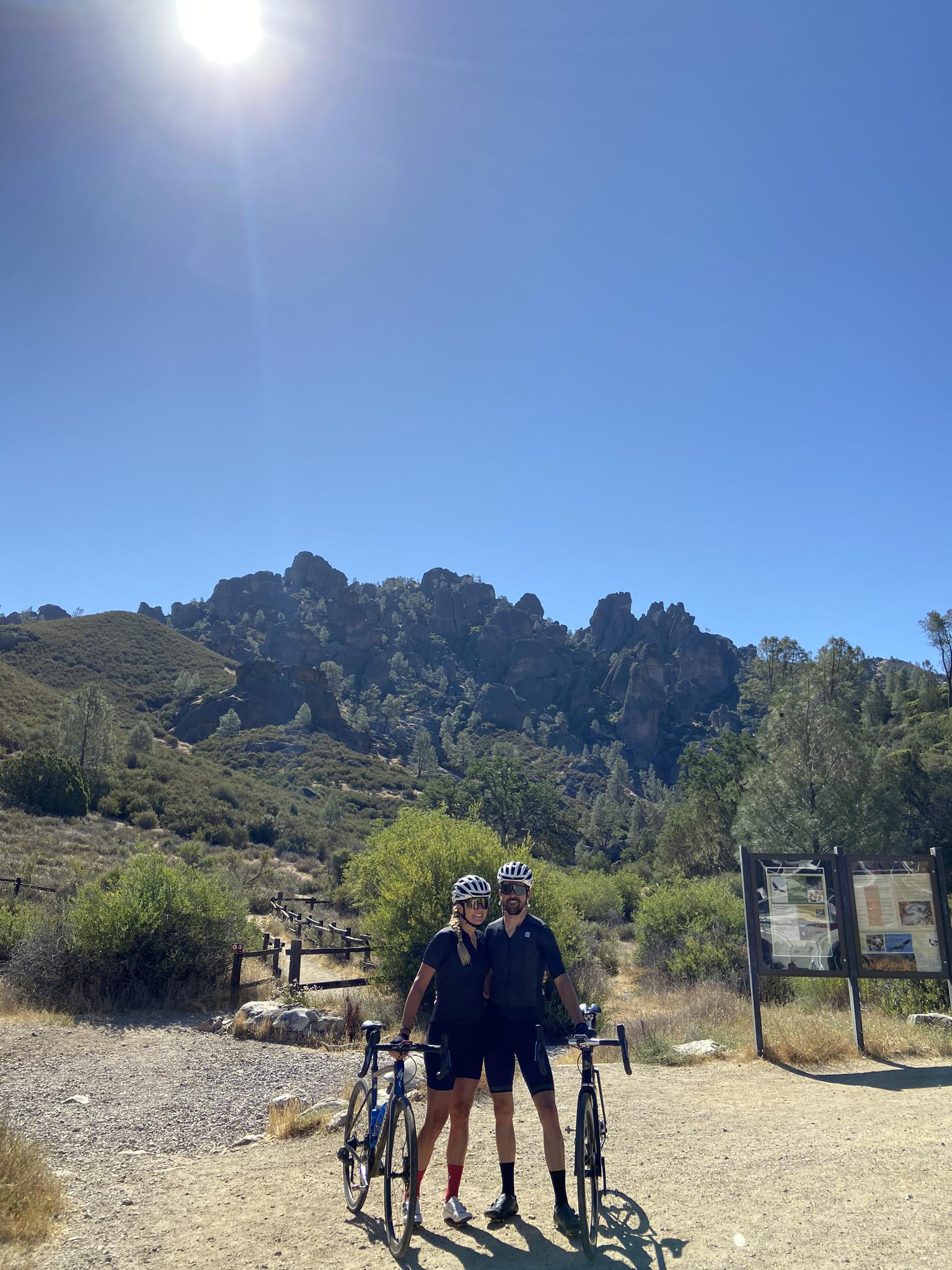 Two cyclists pose with their bikes on a sunny day with rocky hills.