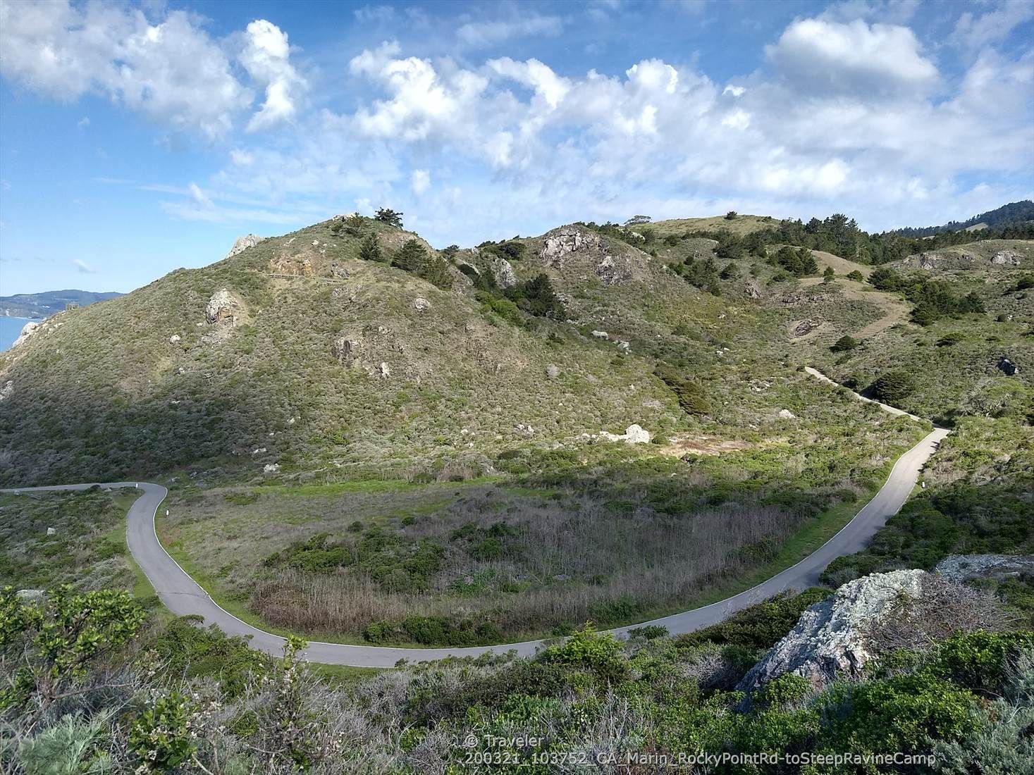 A winding road curves through green, rocky hills under a partly cloudy sky. - Marin Coastal Bike Route