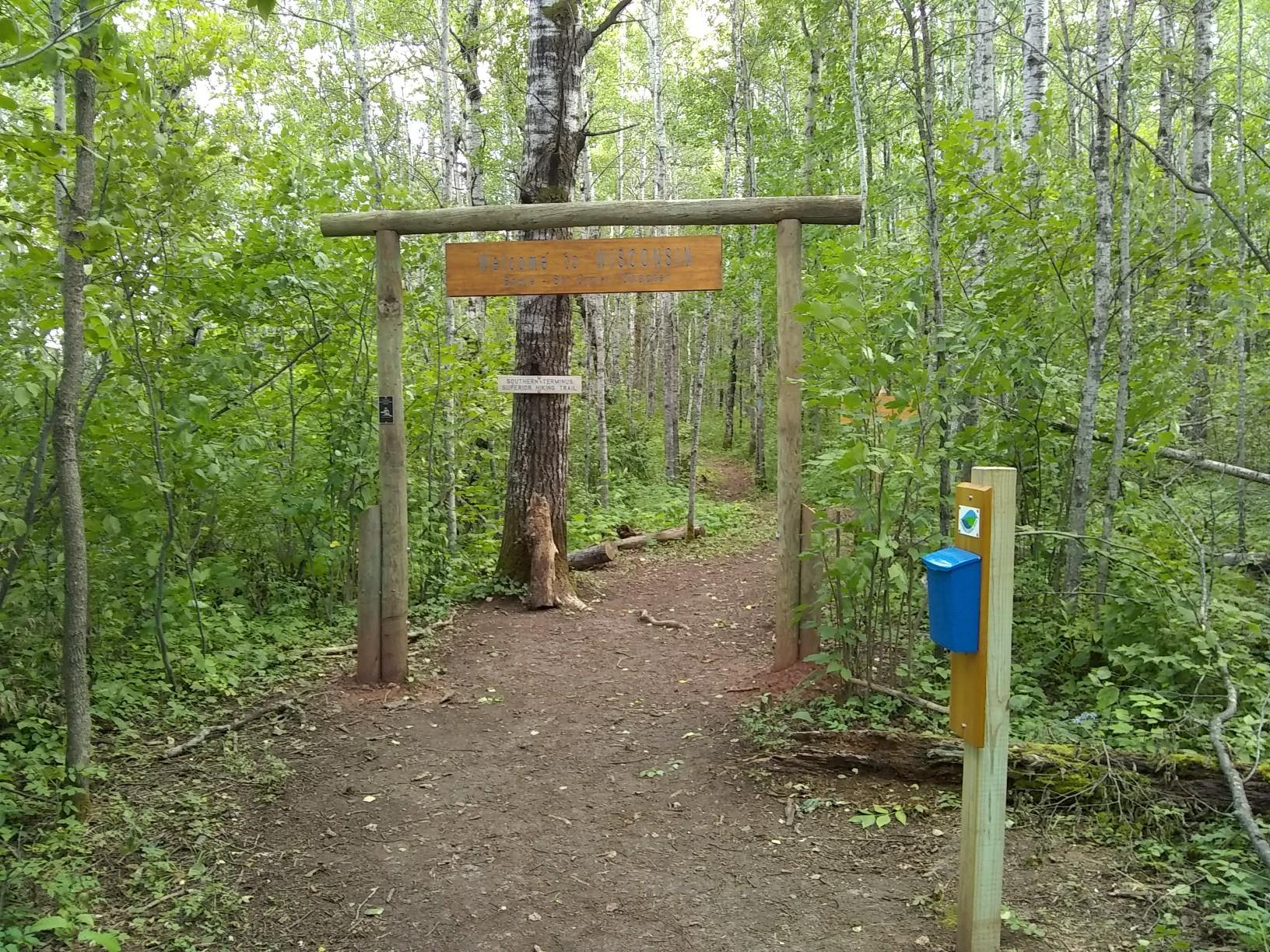A wooden archway marks the entrance to a dirt path in a green forest.