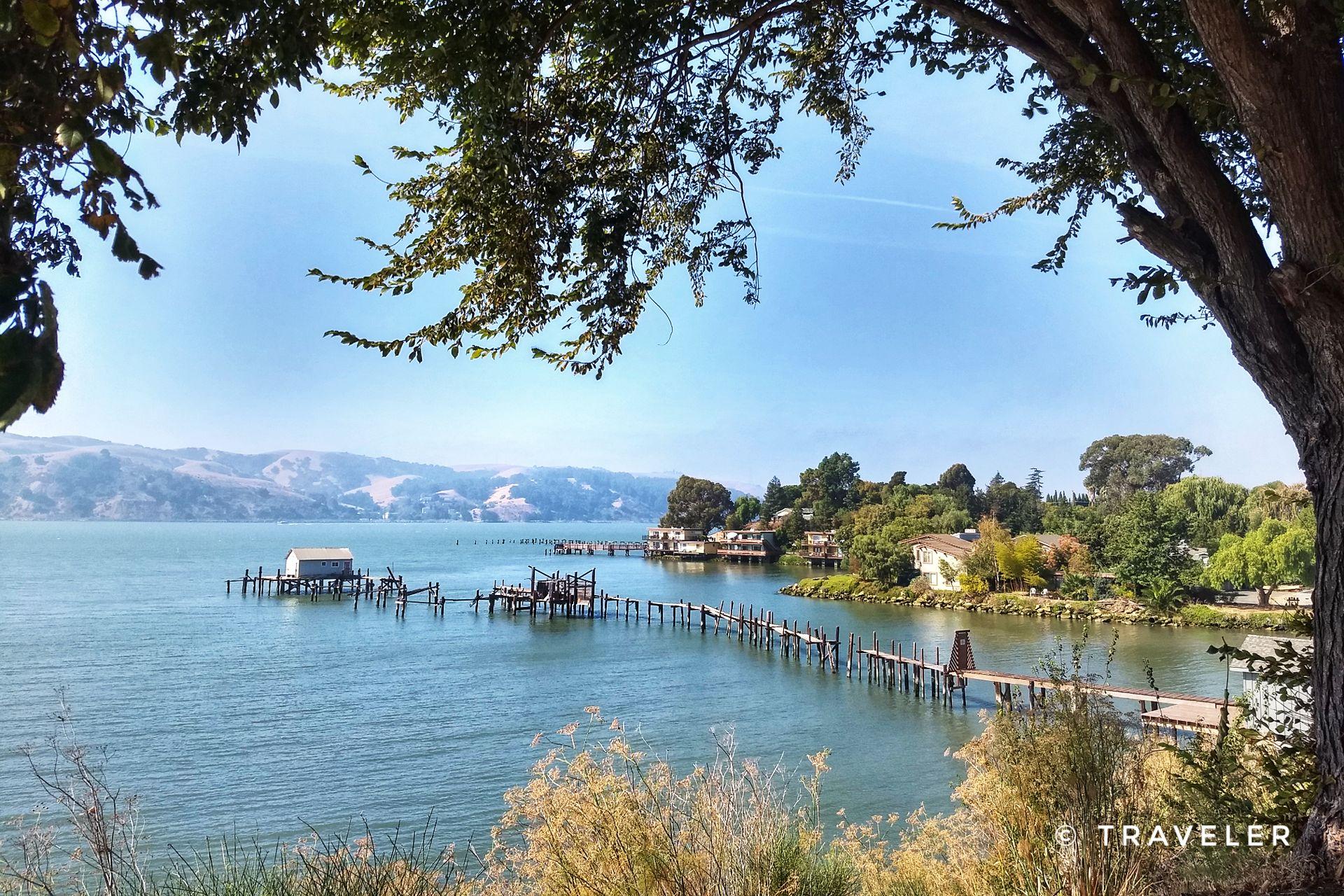 Serene water scene with houses on stilts and a long pier, framed by trees.
