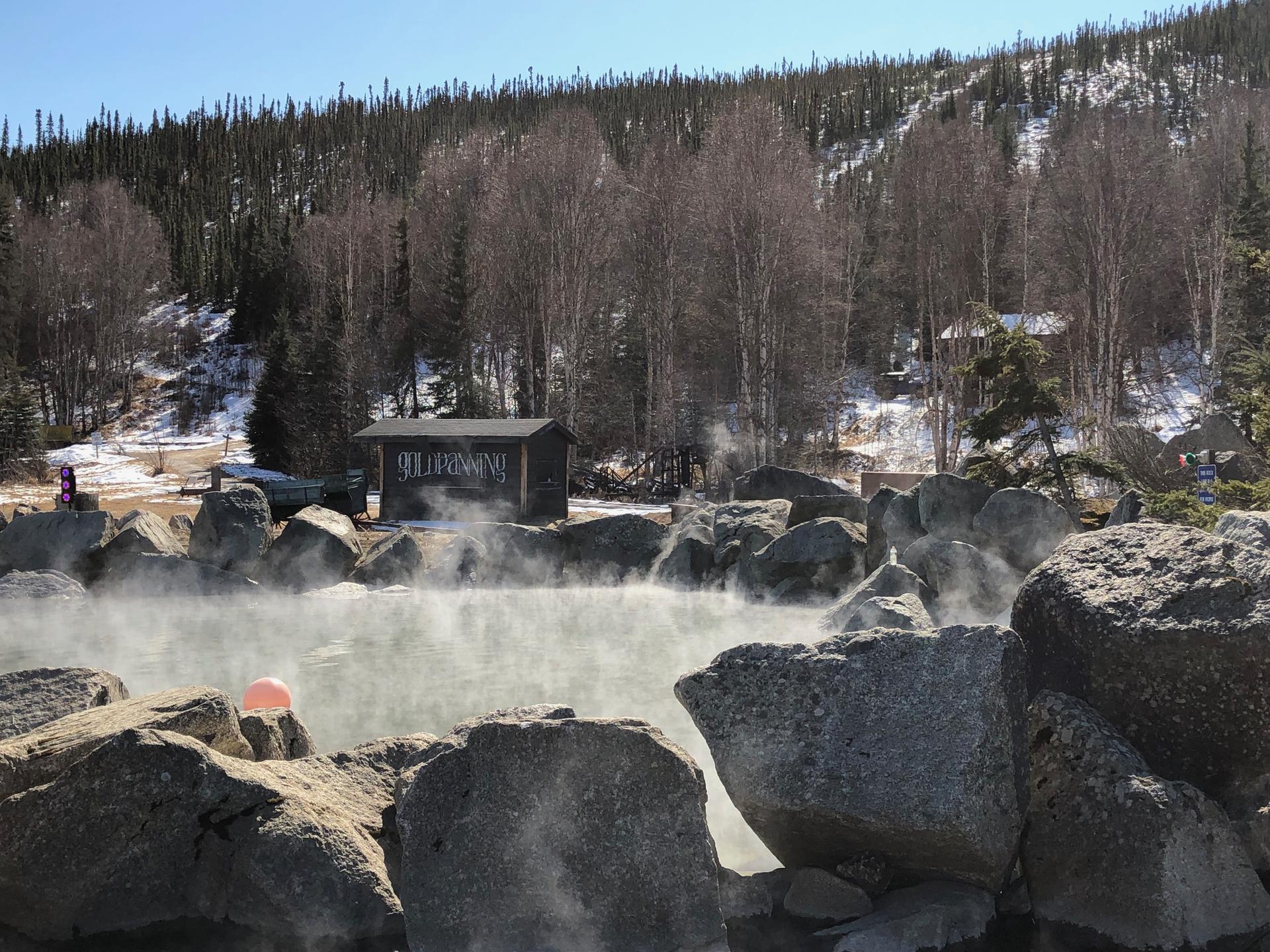 Steaming hot spring pool surrounded by rocks, with snowy hills and trees.