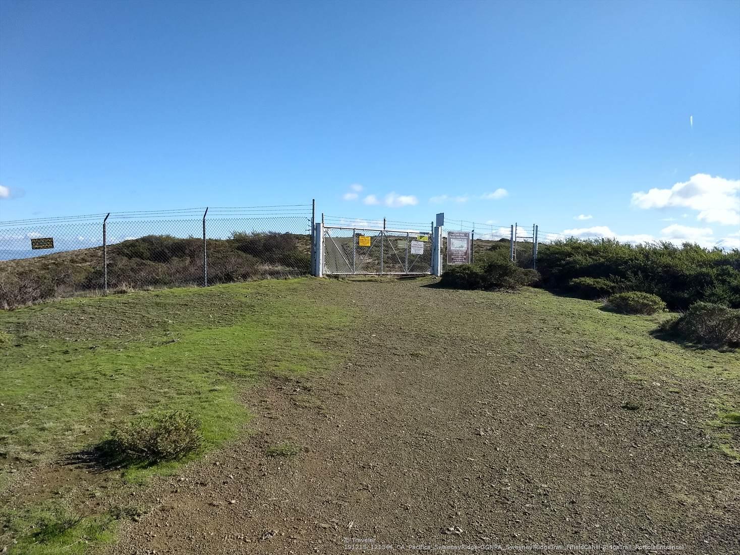 A chain-link fence and gate block a dirt path on a sunny hill.