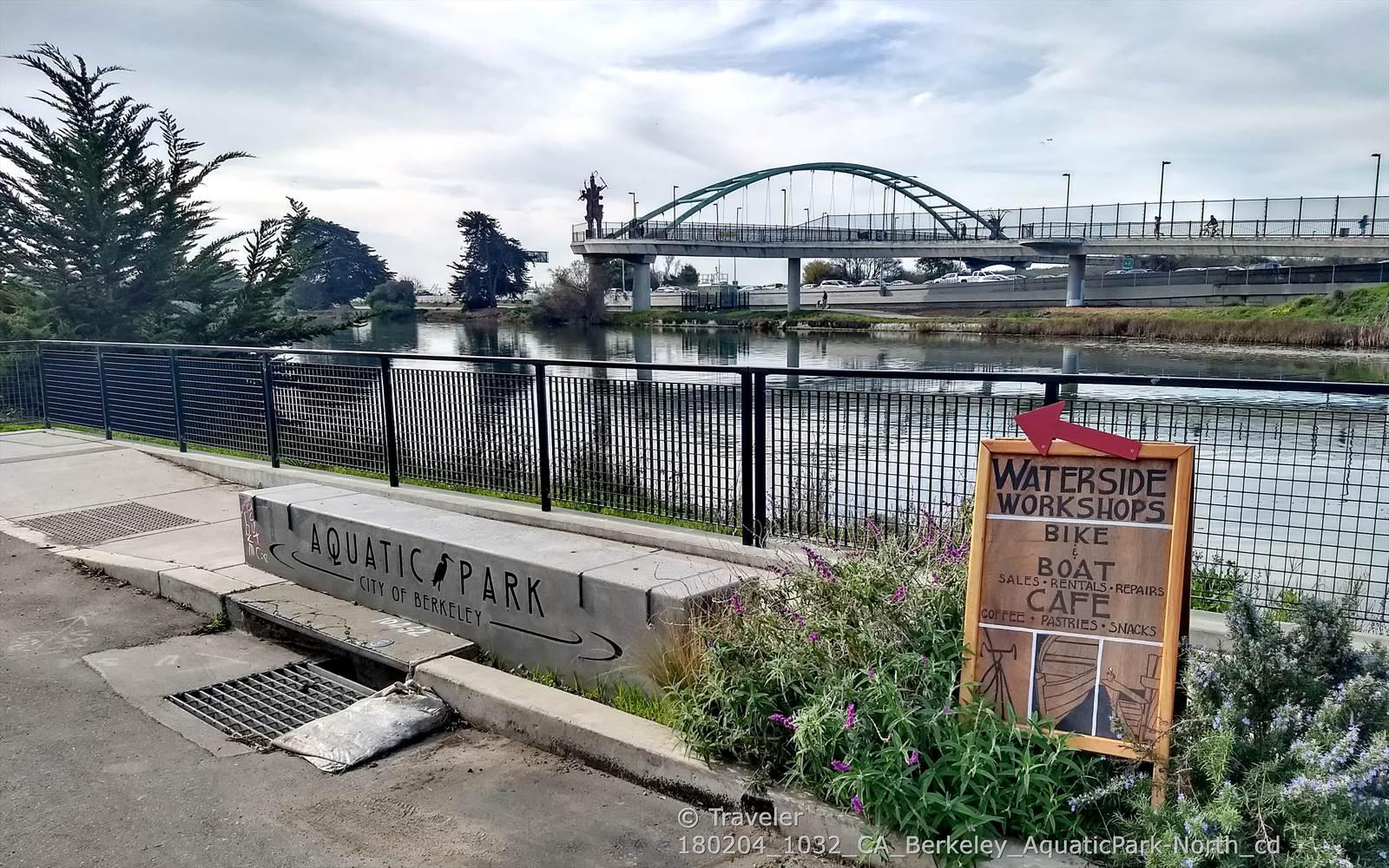 Waterside park with a sign, concrete bench, fence, and bridge under cloudy sky.