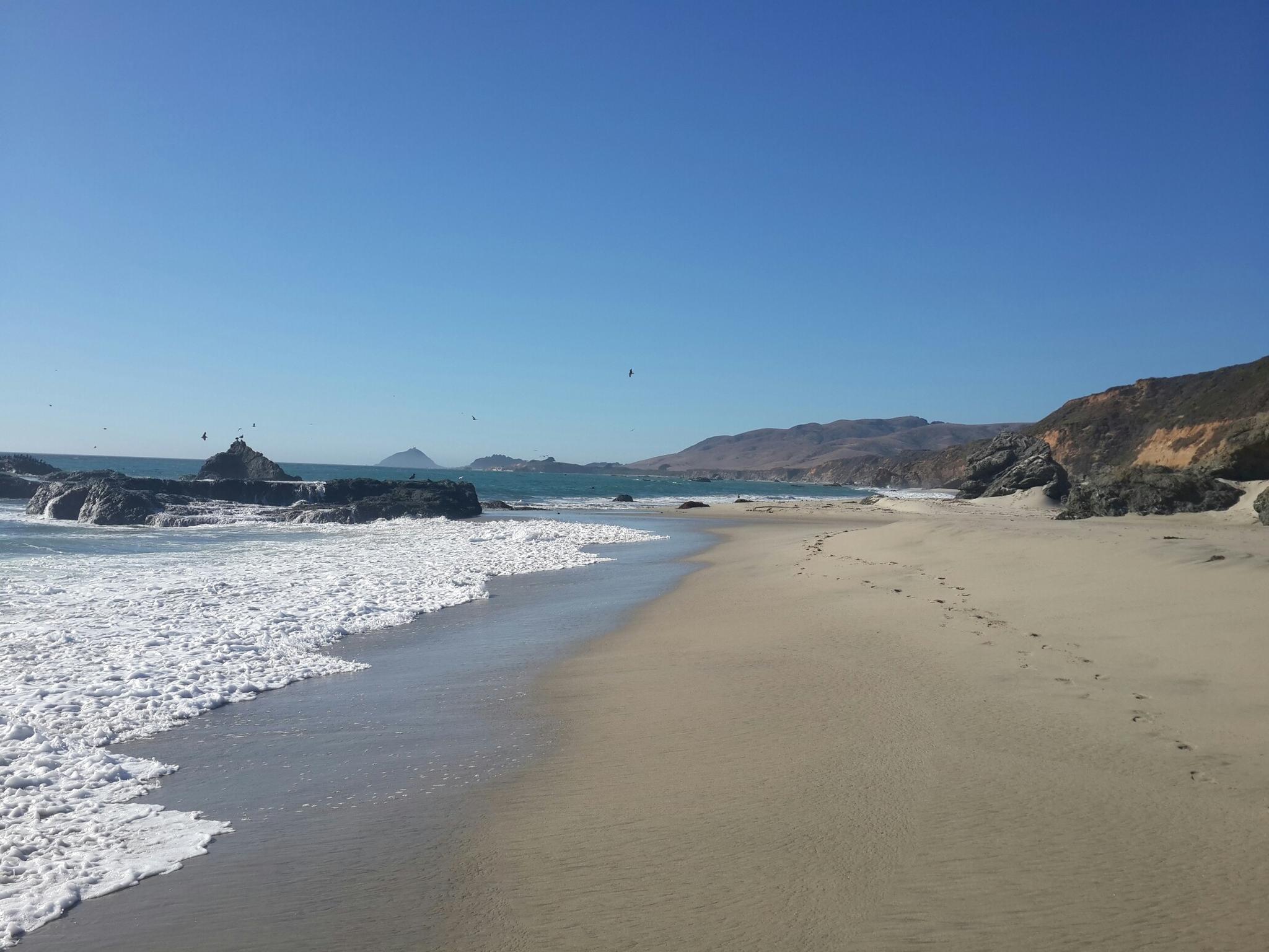 Sunny sandy beach with crashing waves, large rocks, and distant hills.