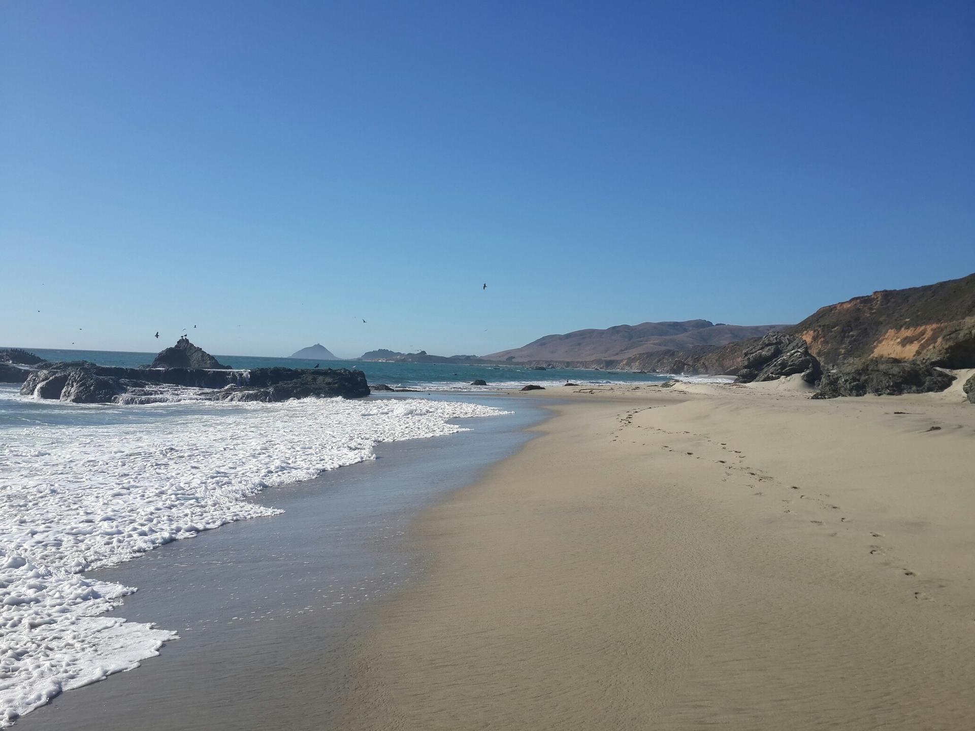 Sunny sandy beach with crashing waves, large rocks, and distant hills.