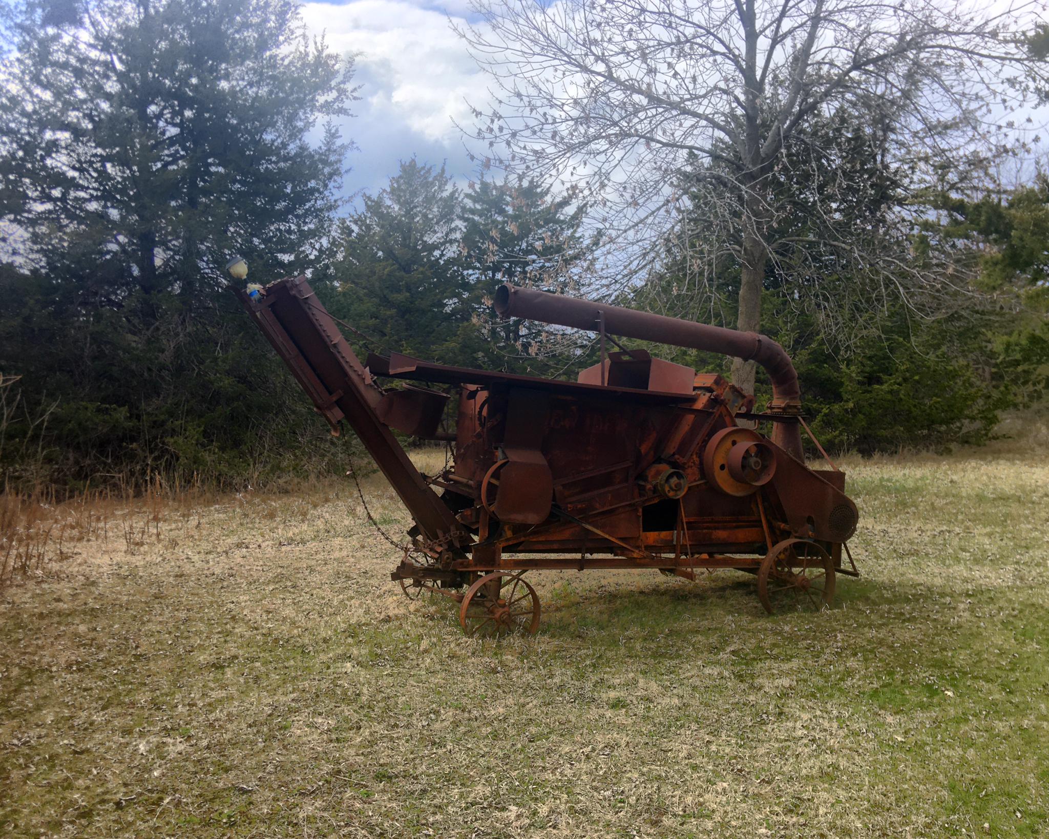 Rusty old farm machinery sits in a grassy field with trees under a cloudy sky.