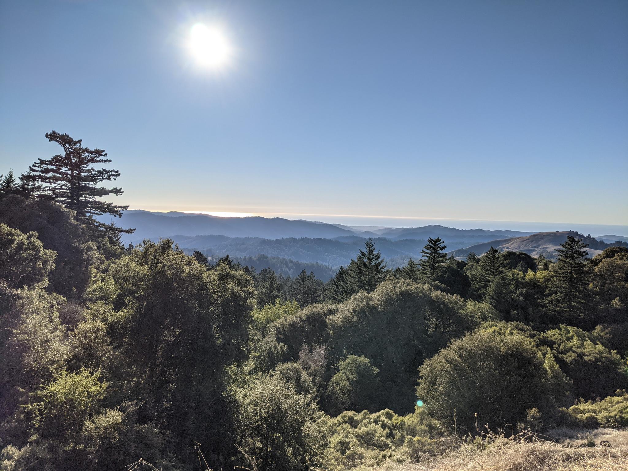 Sunny mountain landscape with forested hills, distant water, and bright sun overhead.