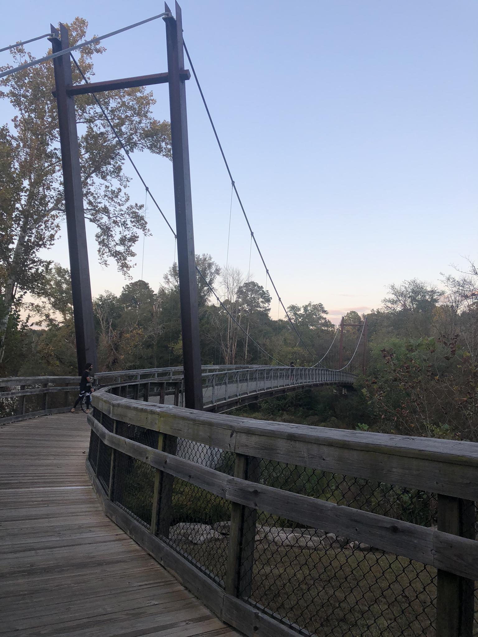 A curved wooden suspension bridge spans over a river with trees.