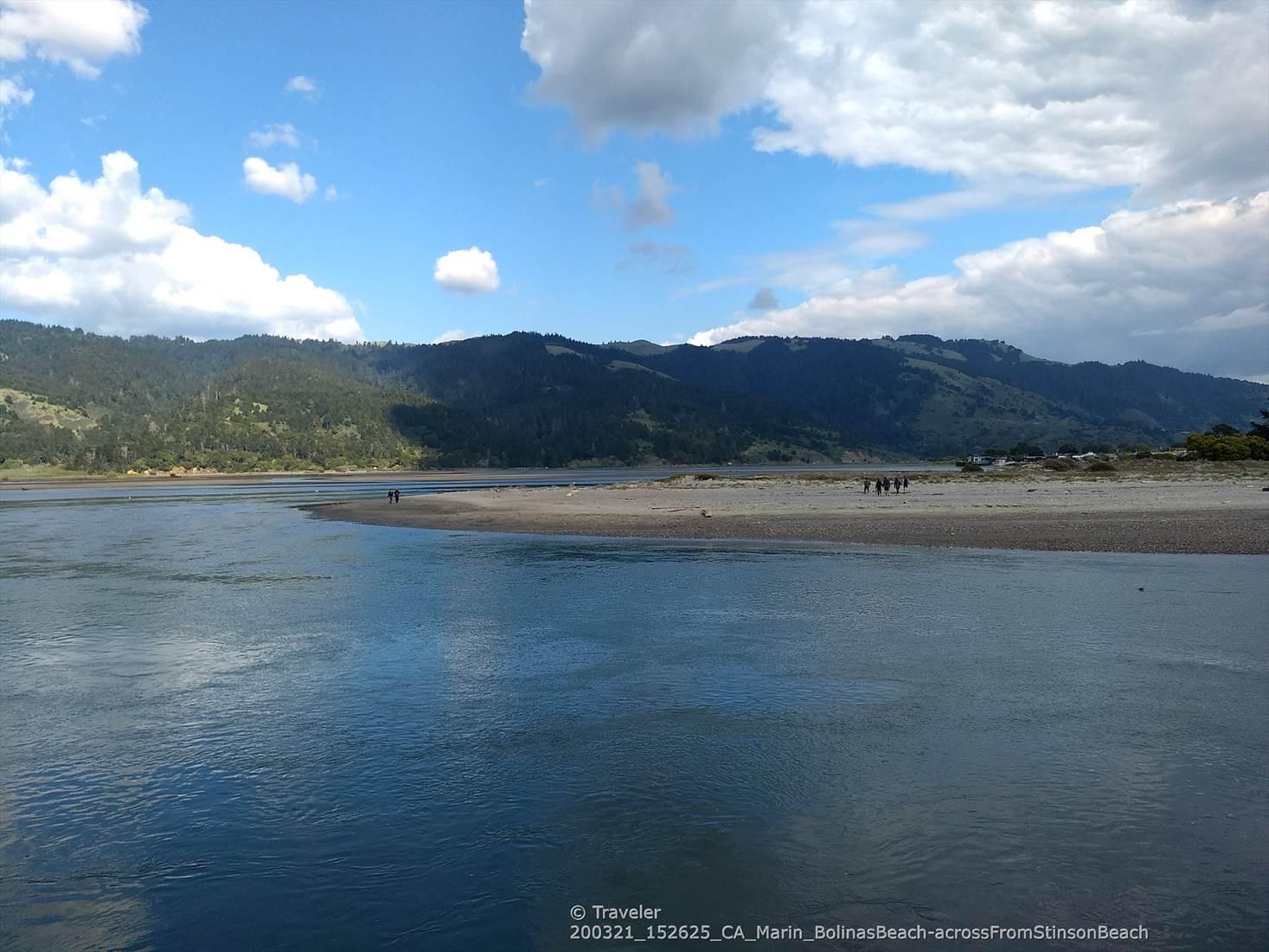 Wide river with a sandy spit and distant forested hills under a blue sky.