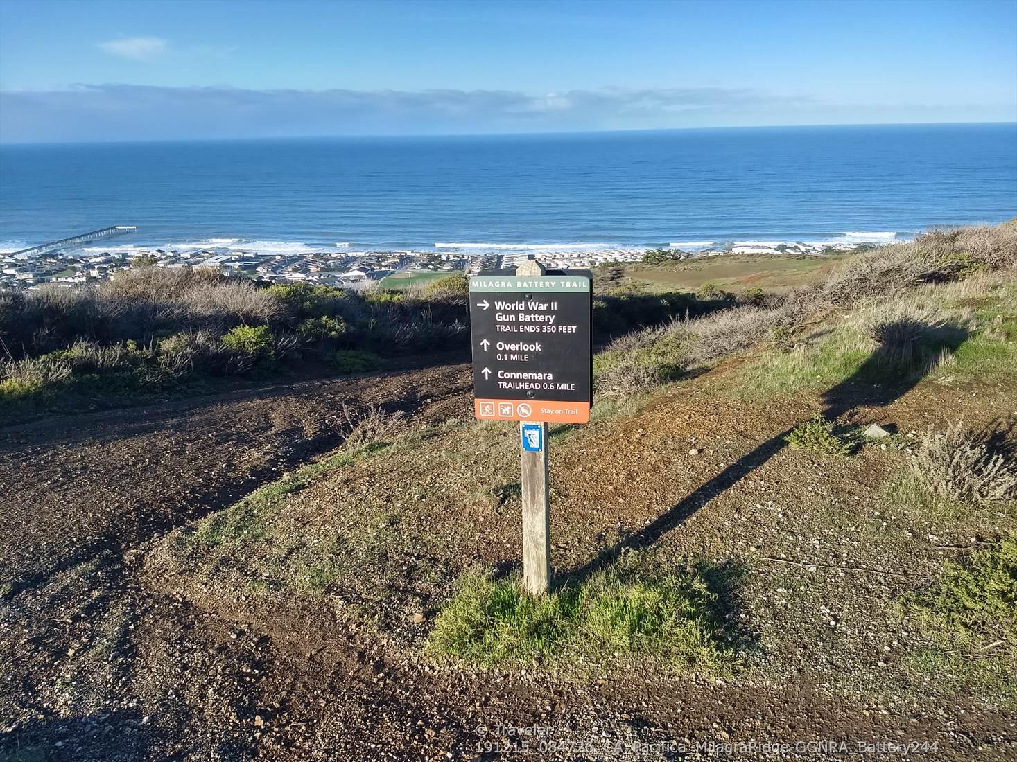 Trail sign on a sunny hillside overlooking the ocean and a distant town.