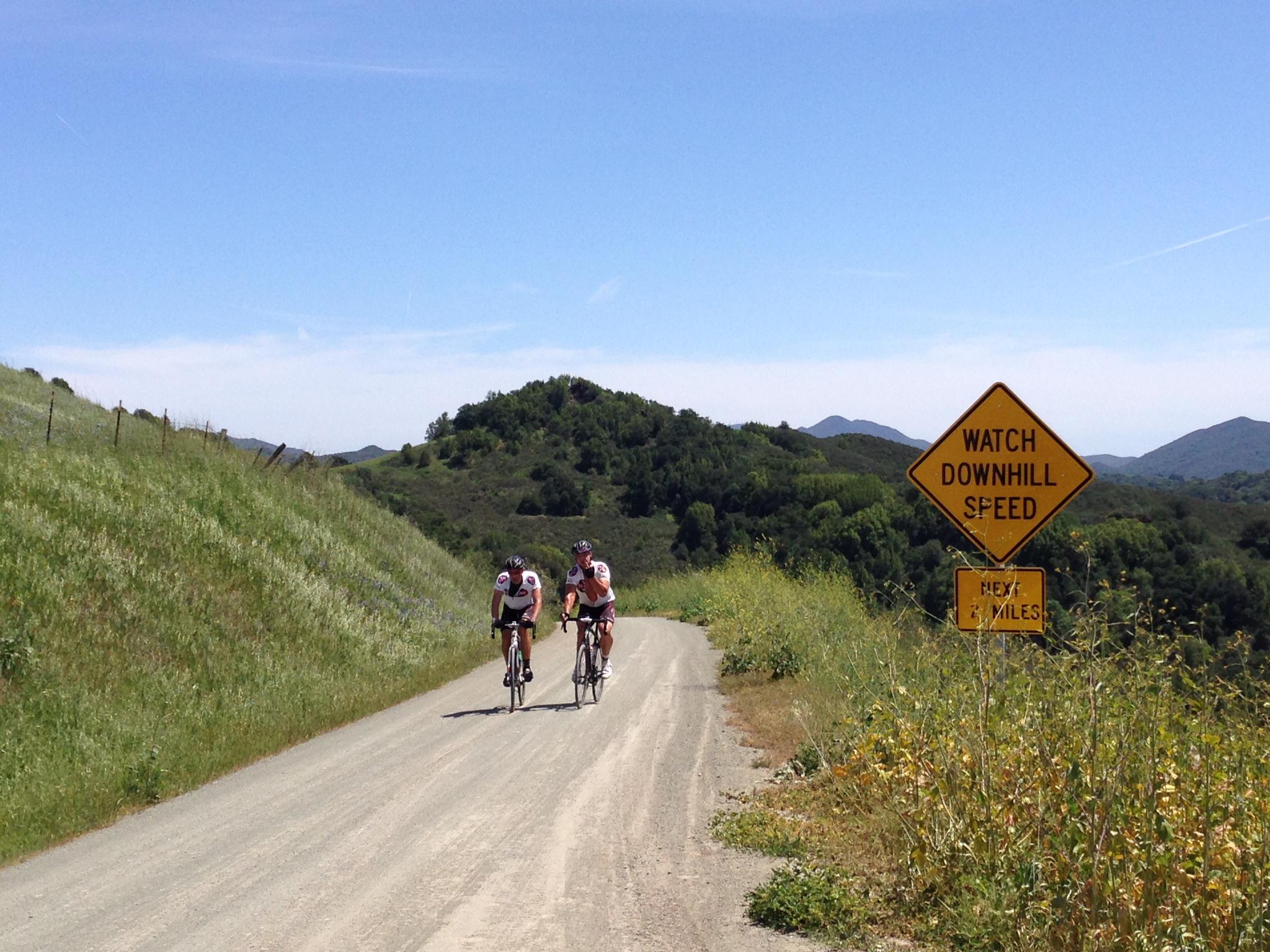Two cyclists ride uphill on a dirt road past a 'Watch Downhill Speed' sign.