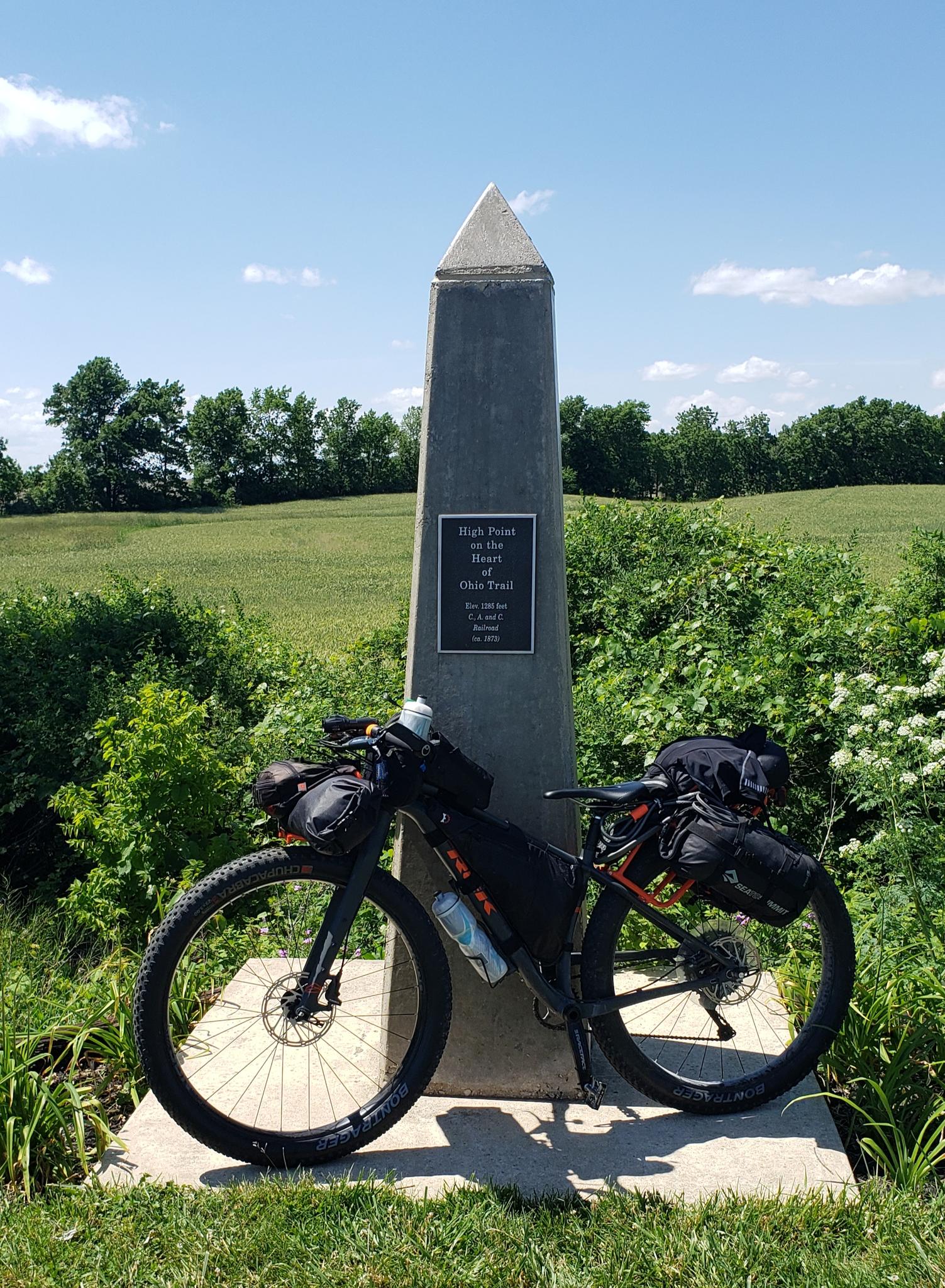 A touring bicycle with bags leans against a gray monument on a sunny day.