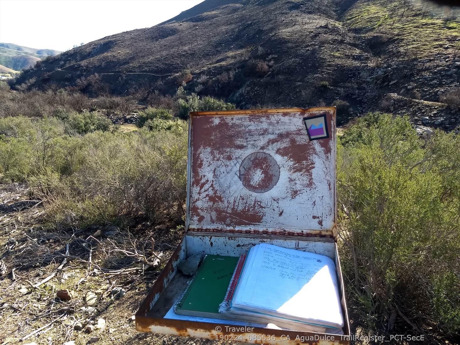 An open, rusty metal box with notebooks sits in dry brush.