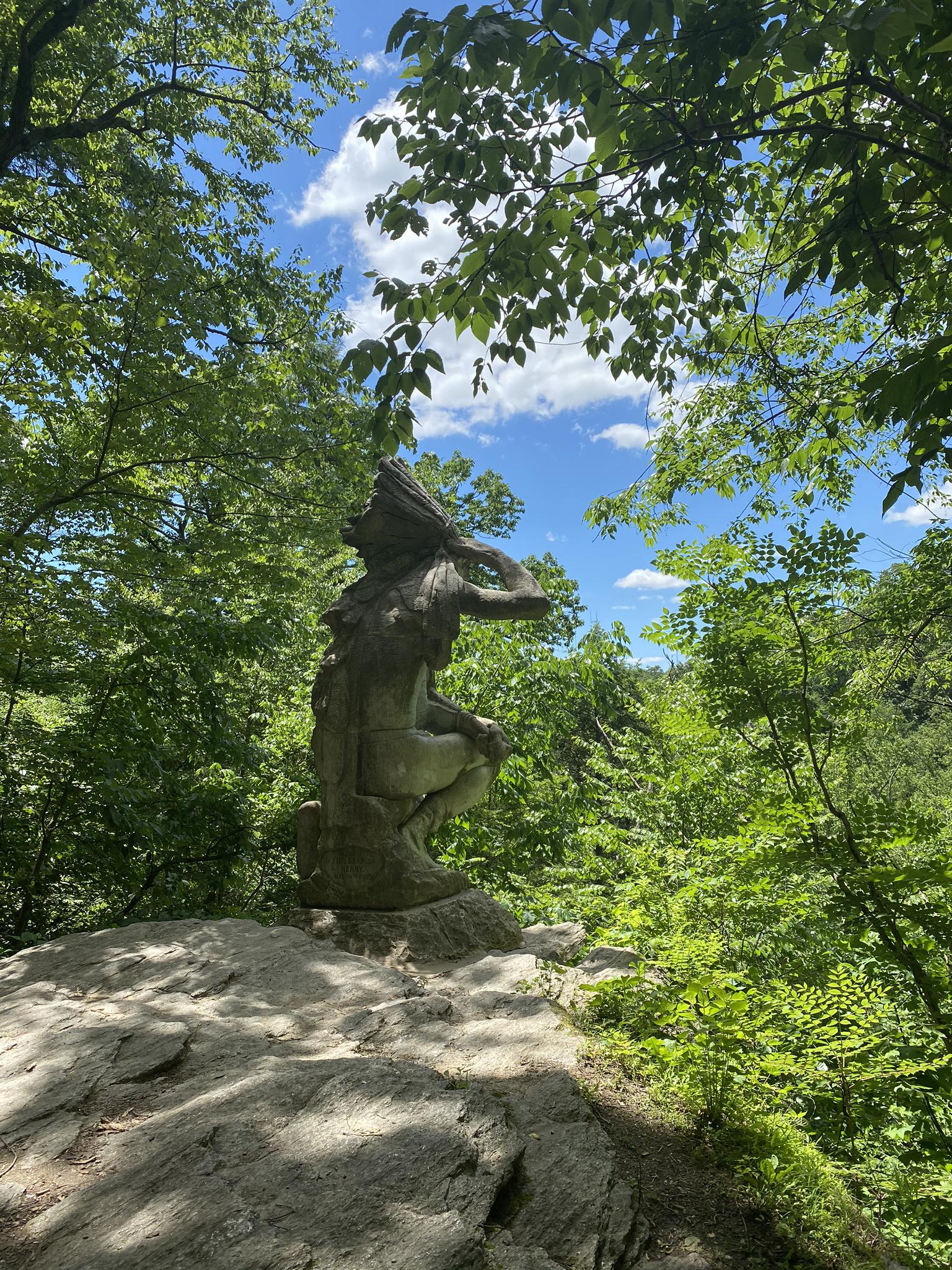 Stone statue on a rock overlooks a vibrant green forest under blue sky.