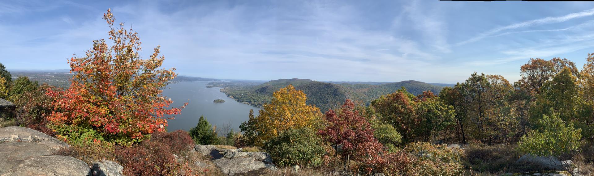 Panoramic autumn view of a lake surrounded by colorful trees and distant hills.