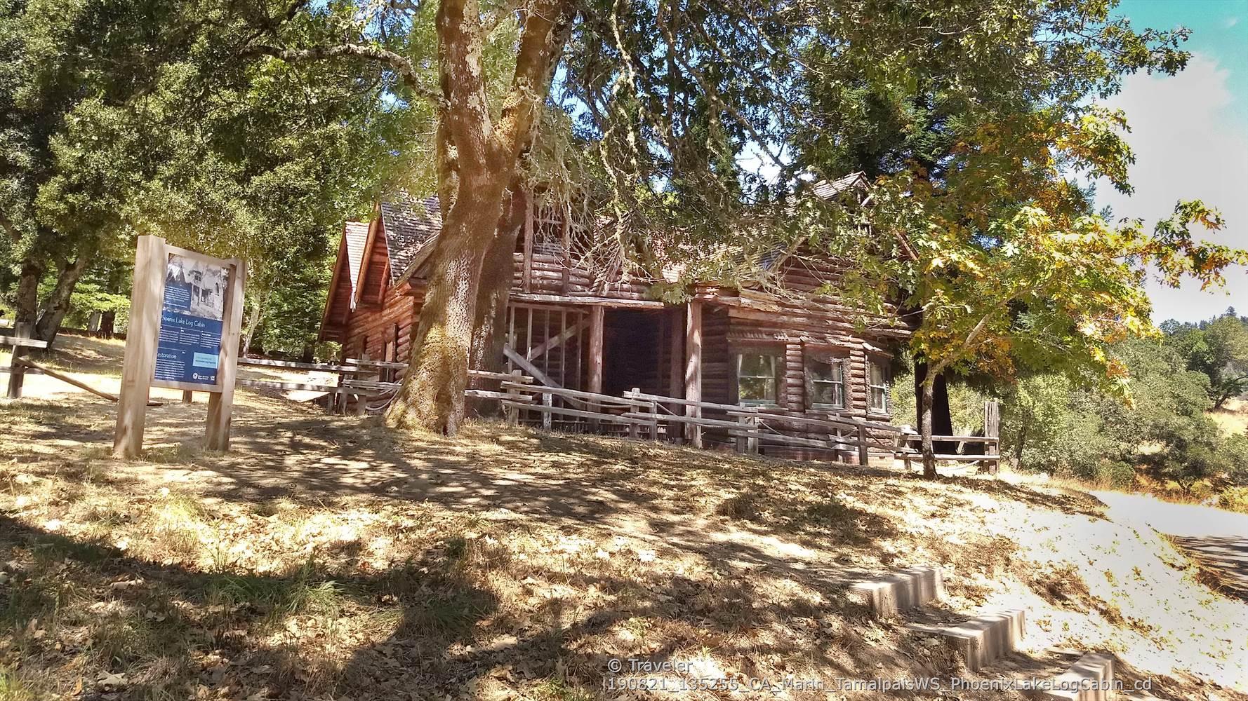 An old, rustic log cabin nestled under large trees on a sunny day.
