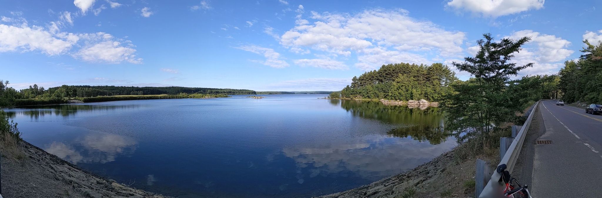 Panoramic view of a wide lake with green shores and a road, sunny day. - Wachusett Reservoir Watershed loop from Worcester