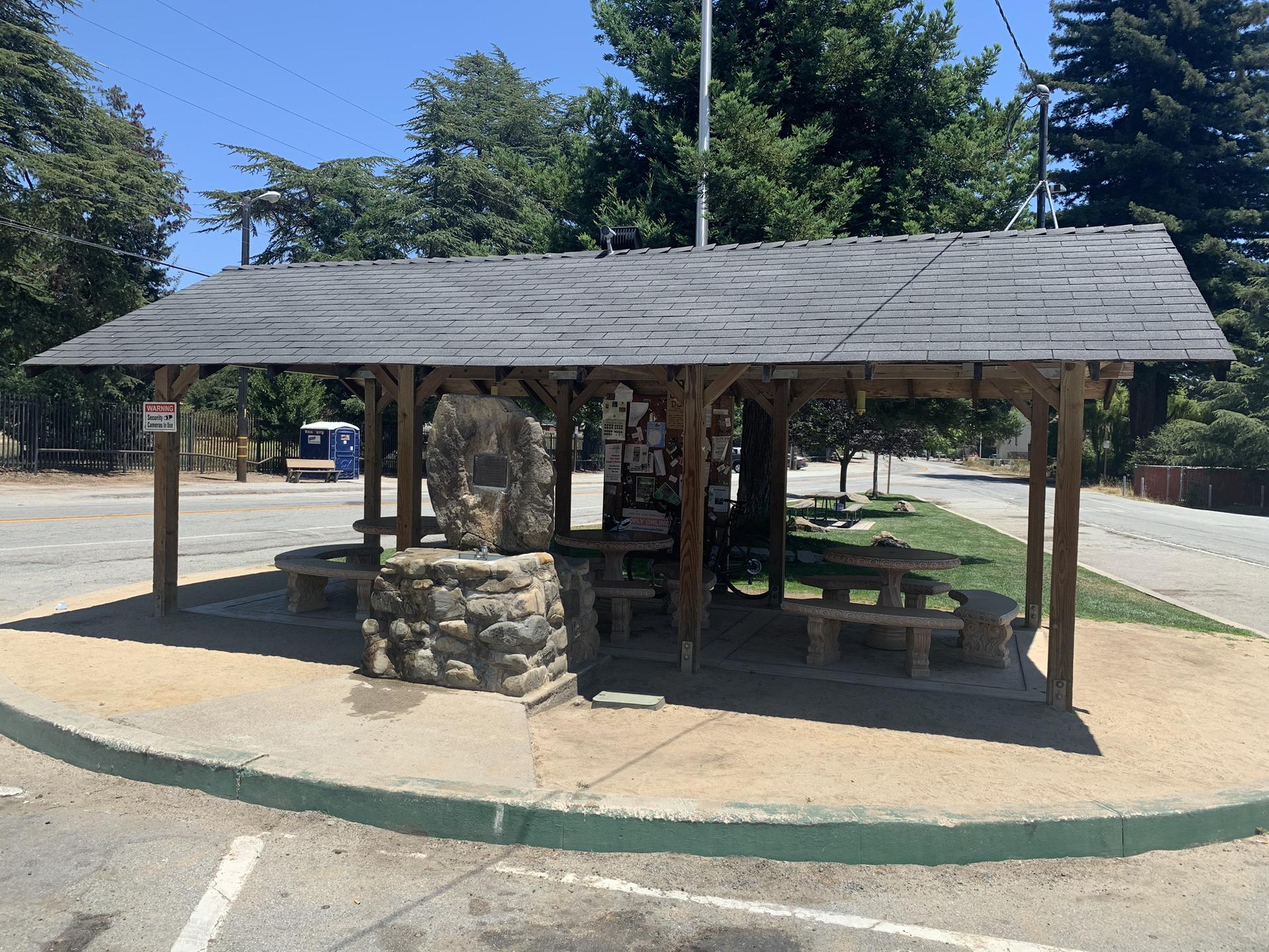 Roadside wooden shelter with stone base and picnic tables on a sunny day.