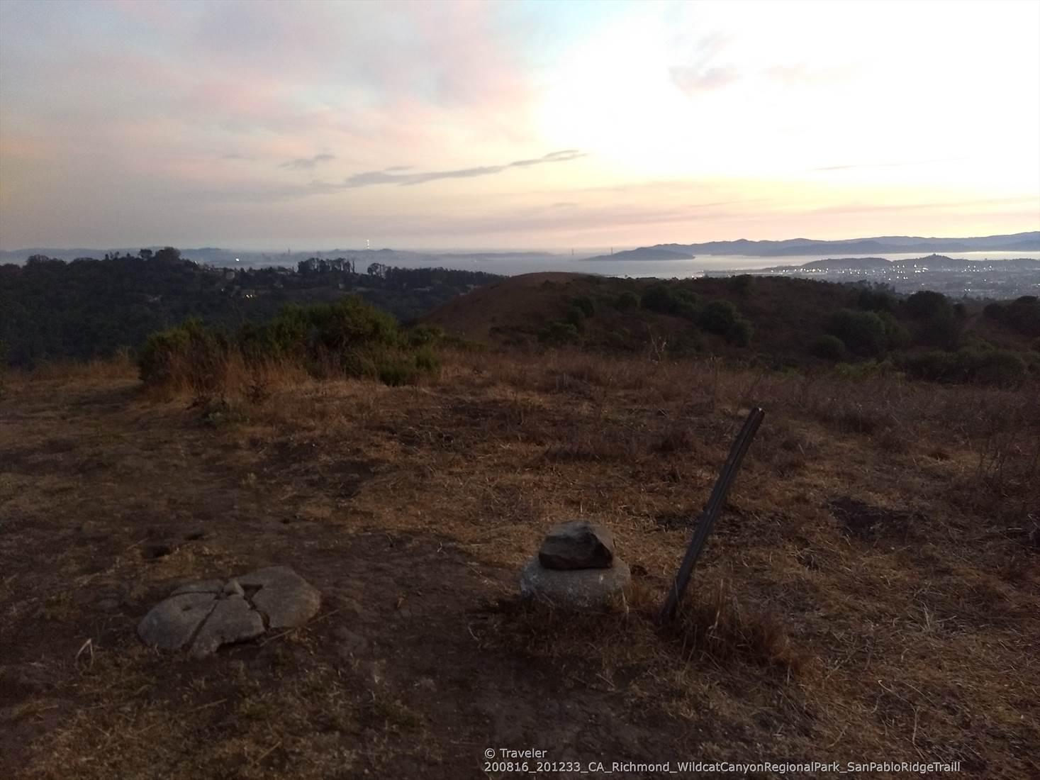 A rocky, dry hilltop overlooks a hazy distant city and body of water.