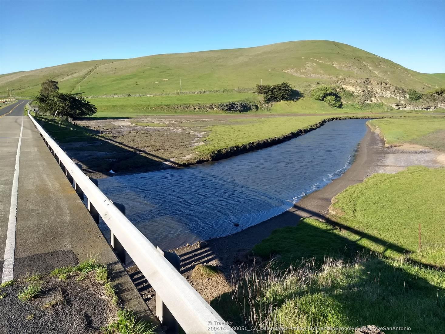 A road with a guardrail next to a narrow river and green hills.