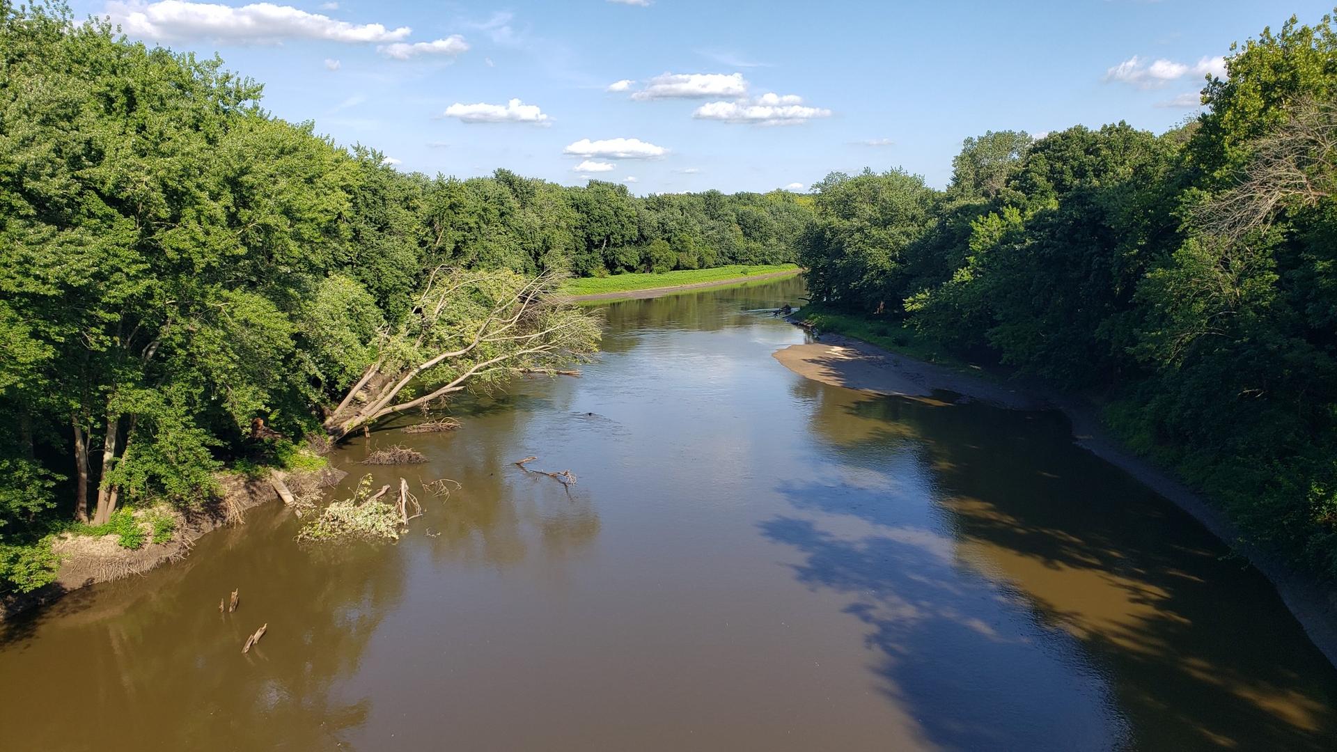 A wide river flows through dense green trees under a bright, cloudy sky.