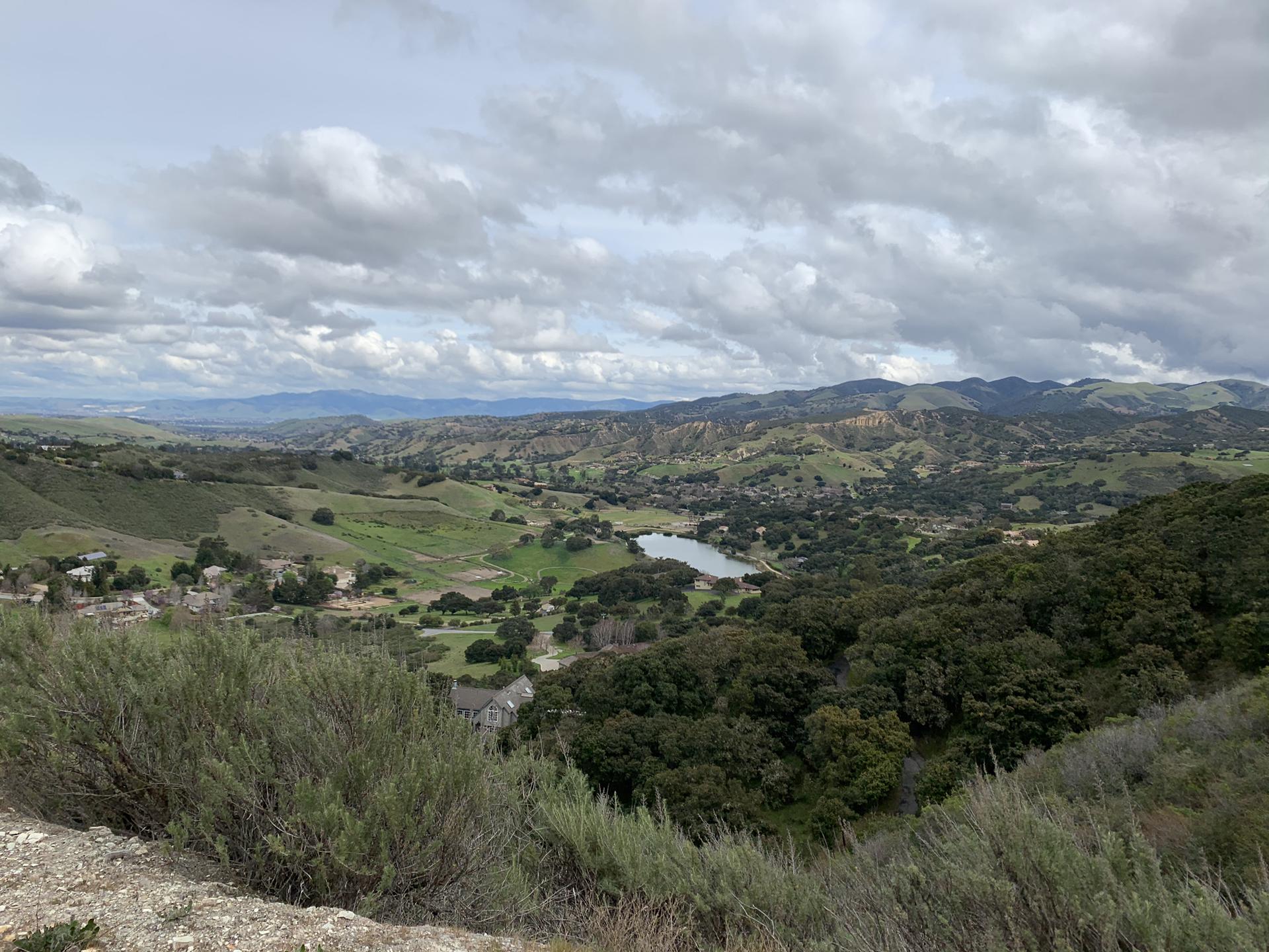 A wide valley with rolling green hills, a lake, under cloudy skies.