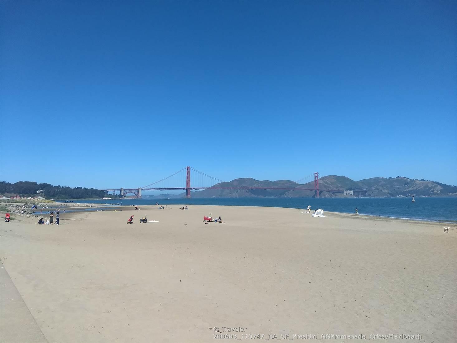 Wide sandy beach with people and a red bridge in the distance under clear sky.