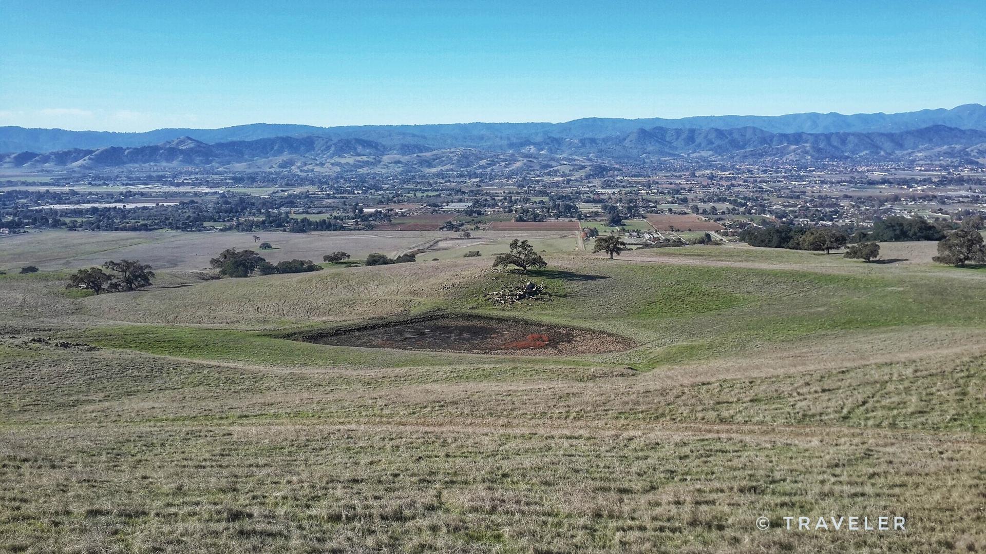 Rolling grassy hills with a small pond, scattered trees, and distant mountains under a clear sky.