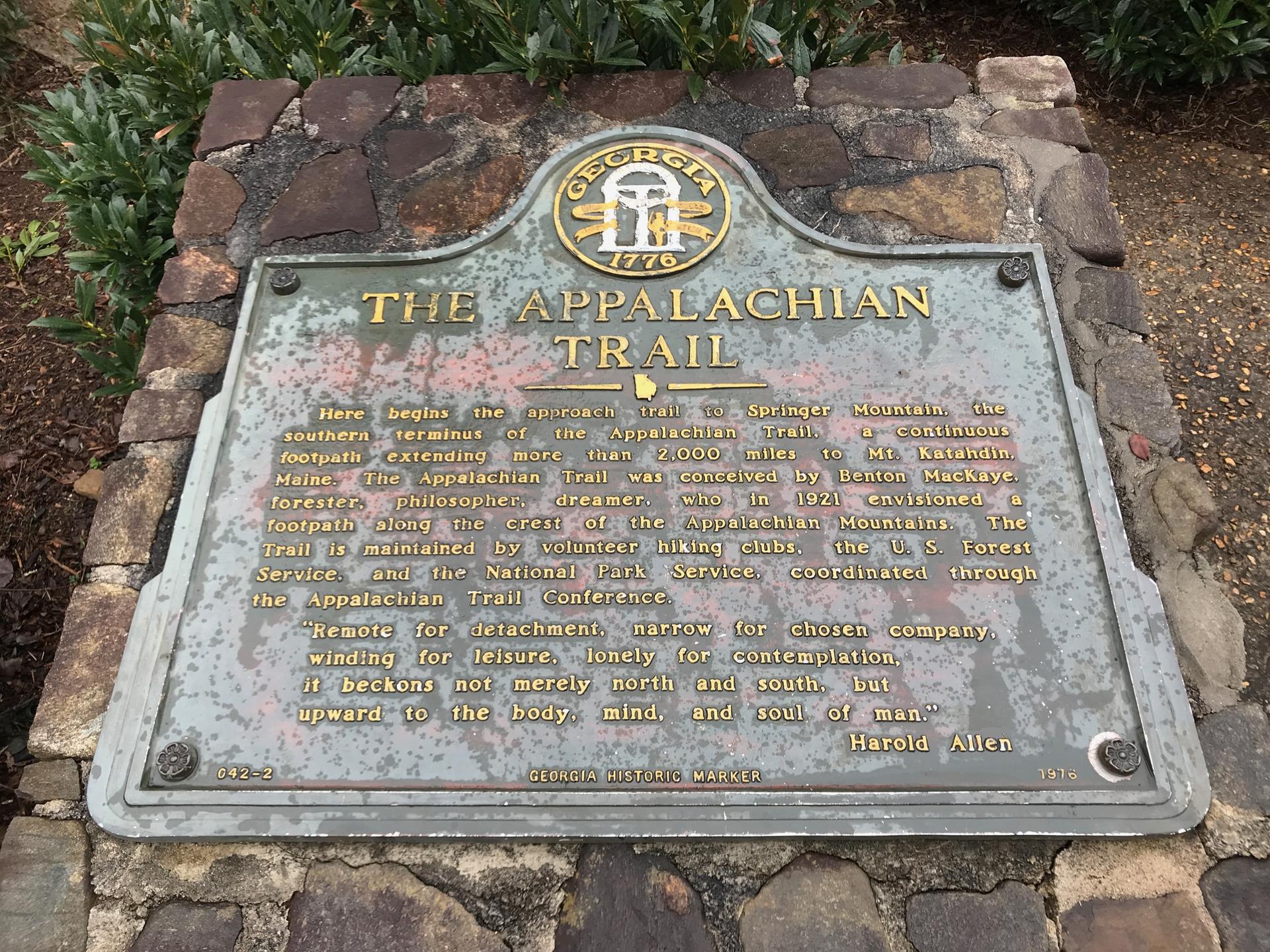 A bronze historical marker on a stone base, surrounded by green plants.
