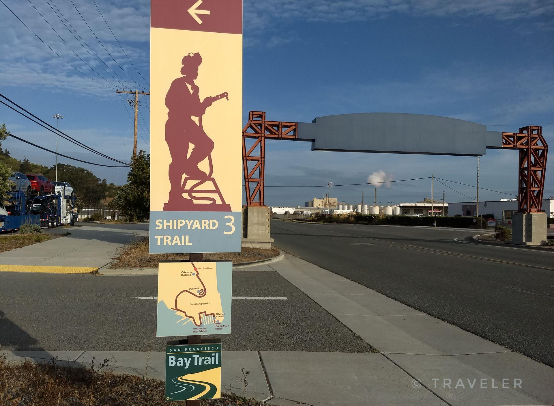 Trail sign with a person silhouette and map, beside a road on a sunny day.