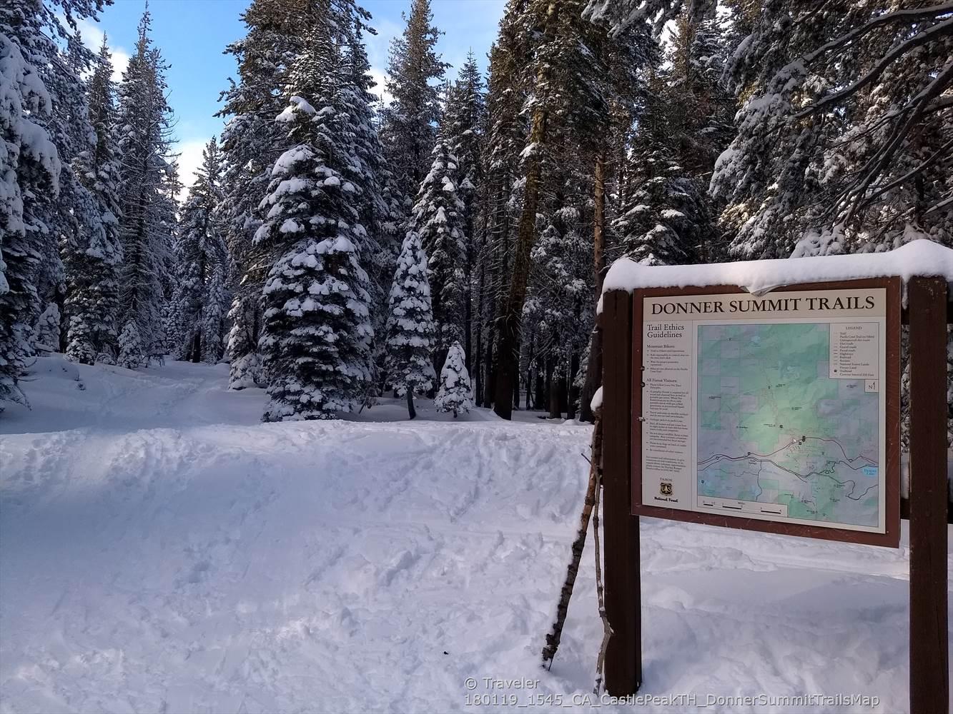 Snow-covered forest path with a trail map sign on a sunny day.