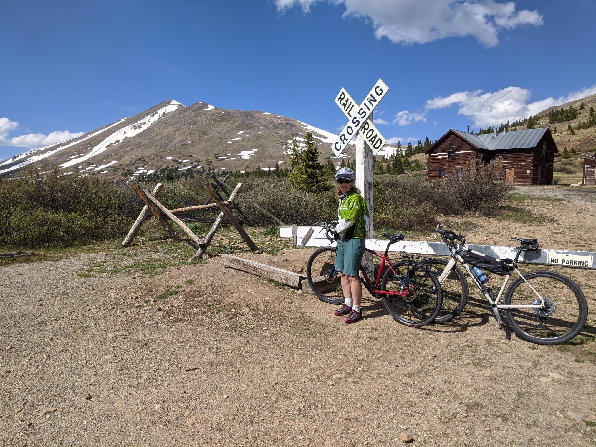 Cyclist stands with two bikes near a railroad sign and snowy mountain.