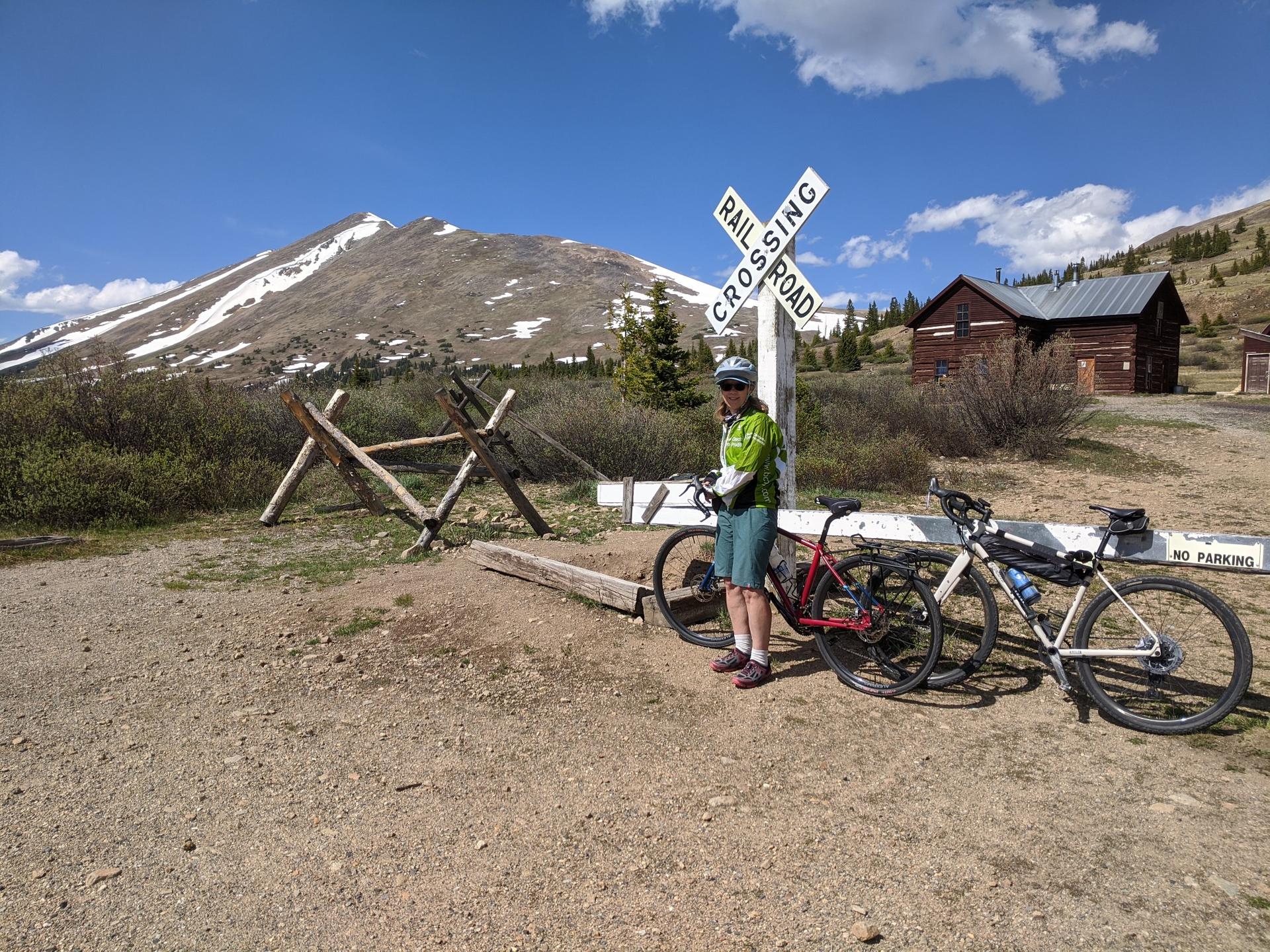 Cyclist stands with two bikes near a railroad sign and snowy mountain.