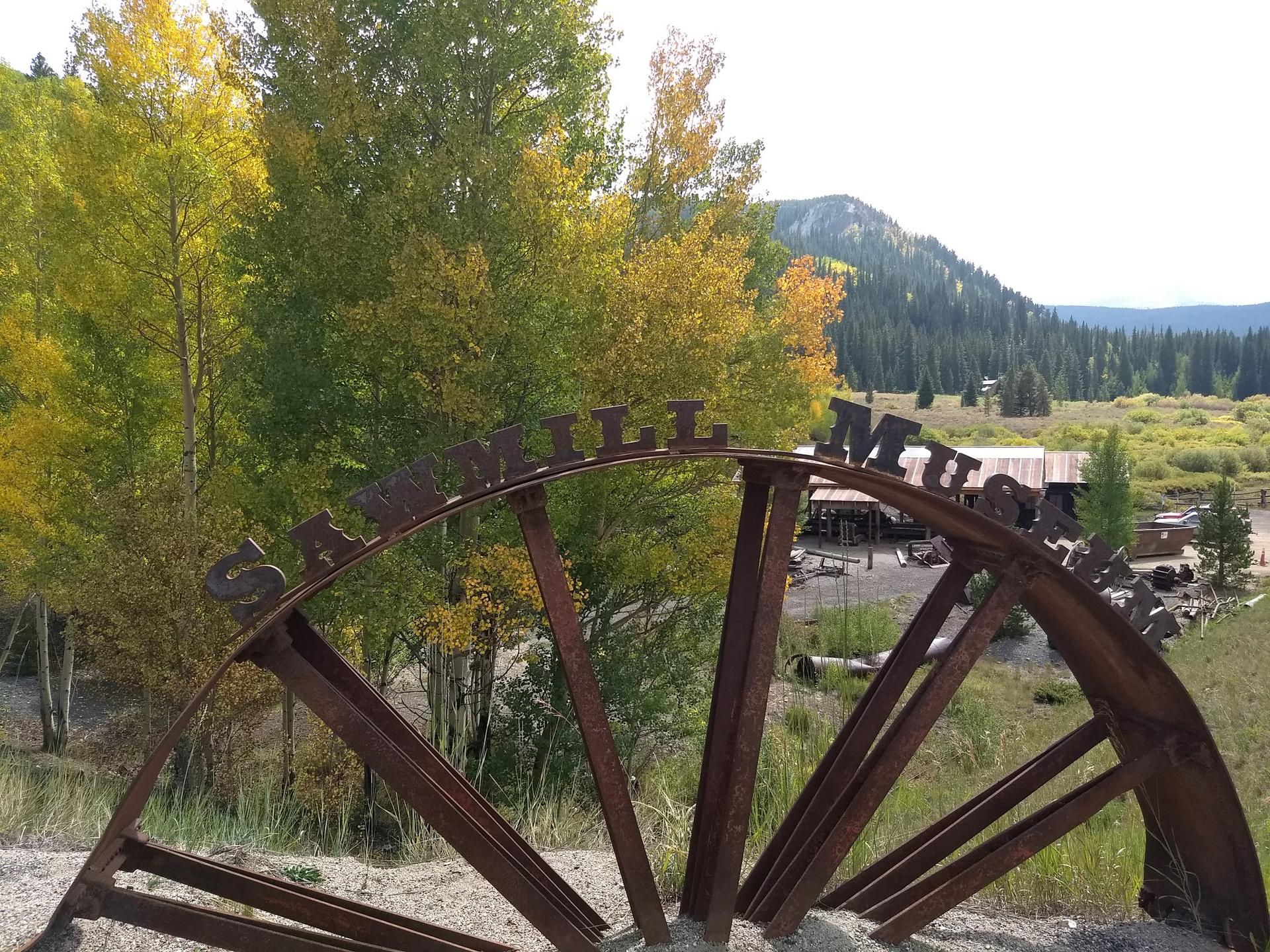 Rusty metal "SAWMILL MUSEUM" sign in front of vibrant autumn trees and hills.
