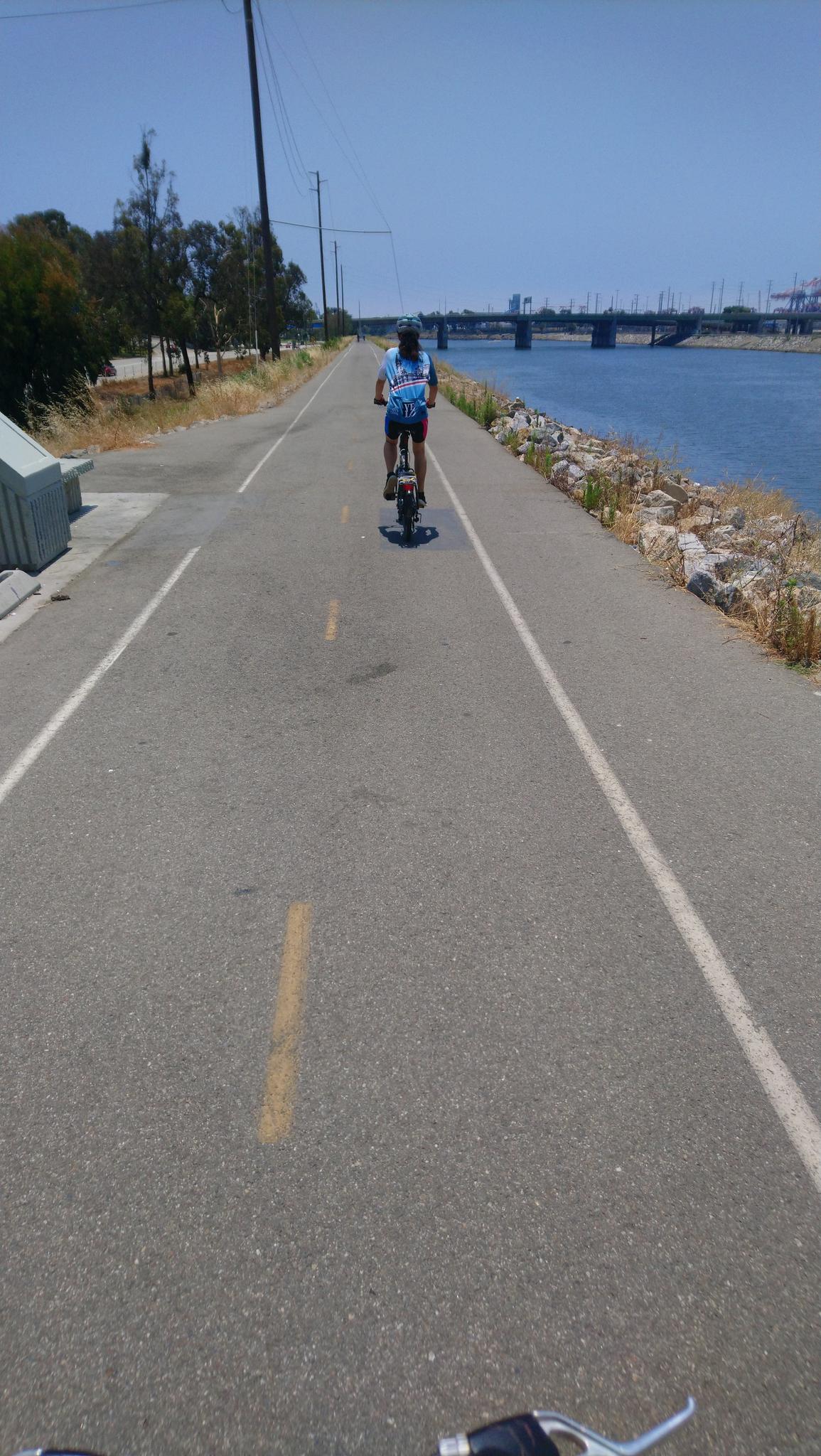 Person on a unicycle rides a paved path beside a river on a sunny day.