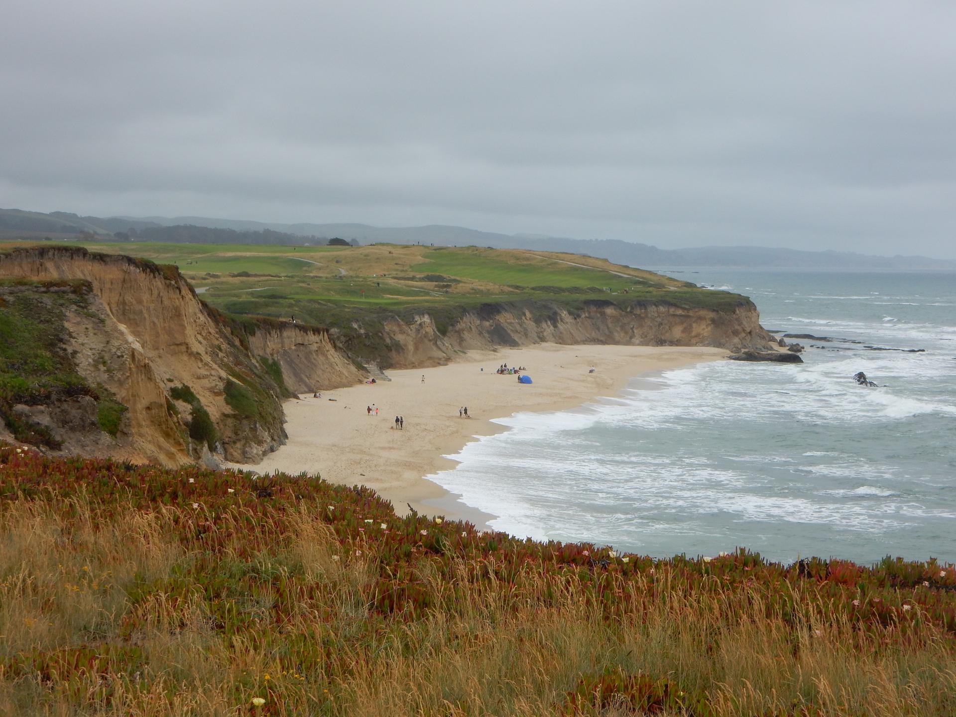Sandy beach with ocean waves, steep cliffs, and distant green hills on a cloudy day.