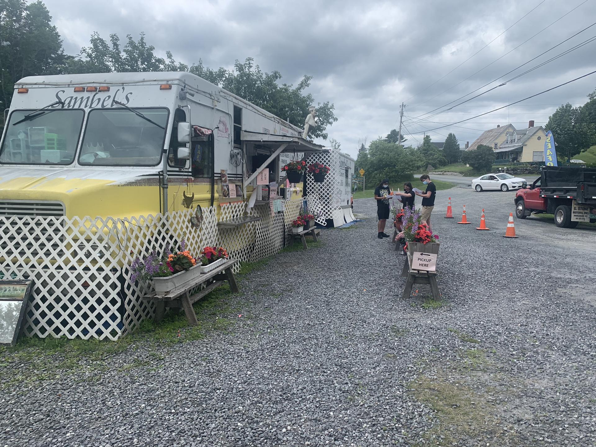 A food truck on gravel, with people standing nearby, under an overcast sky.