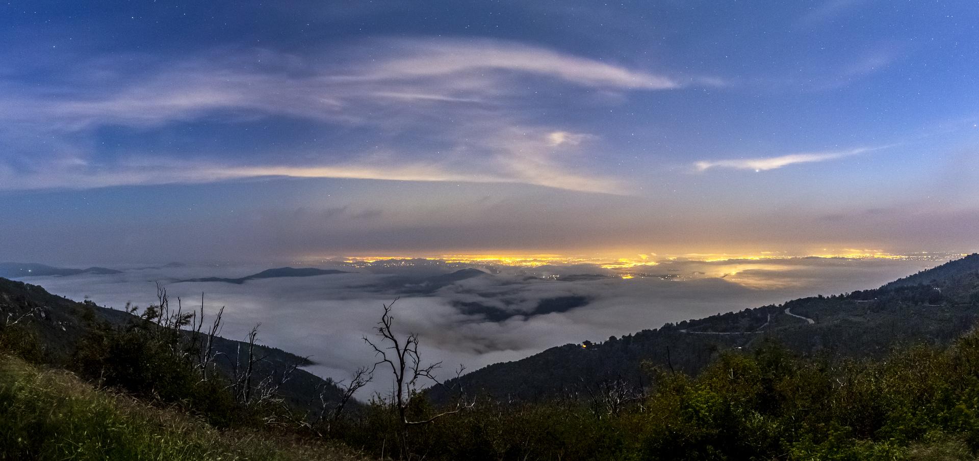 Panoramic mountain view at twilight with glowing city lights under a cloudy sky.
