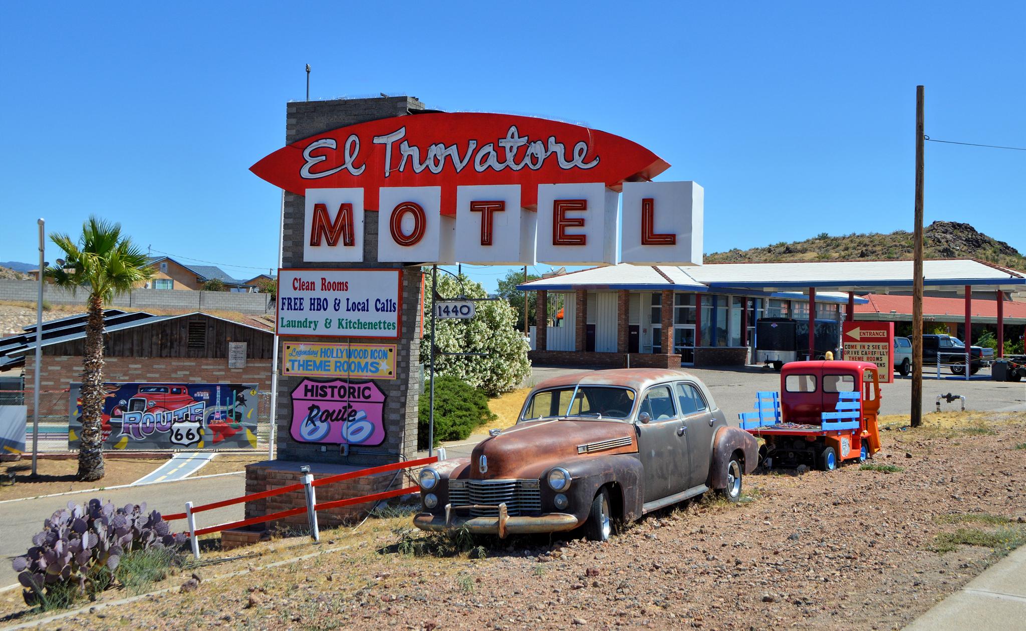 A rusty vintage car parked by a large motel sign on a sunny day.