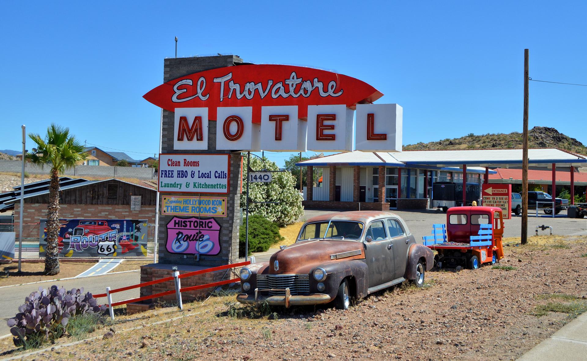 A rusty vintage car parked by a large motel sign on a sunny day.