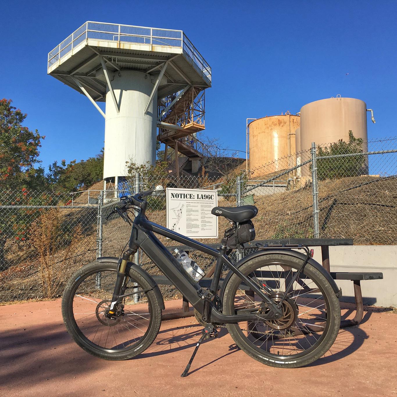 Black bicycle parked on dirt near industrial towers under a clear blue sky.
