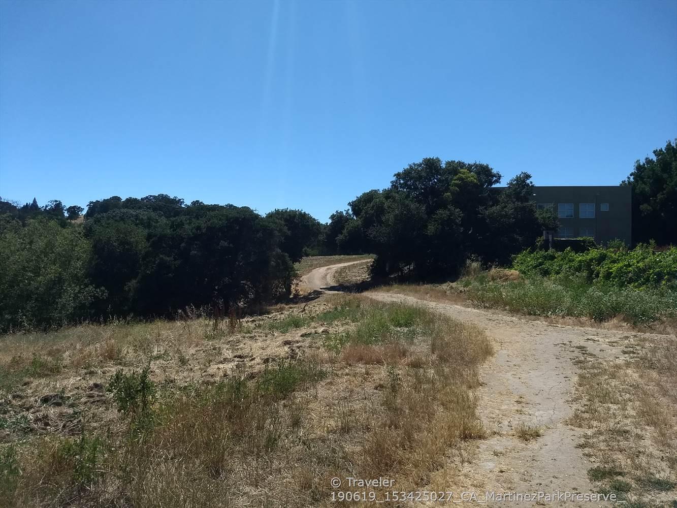 Winding dirt path through dry fields and green trees under clear sky.