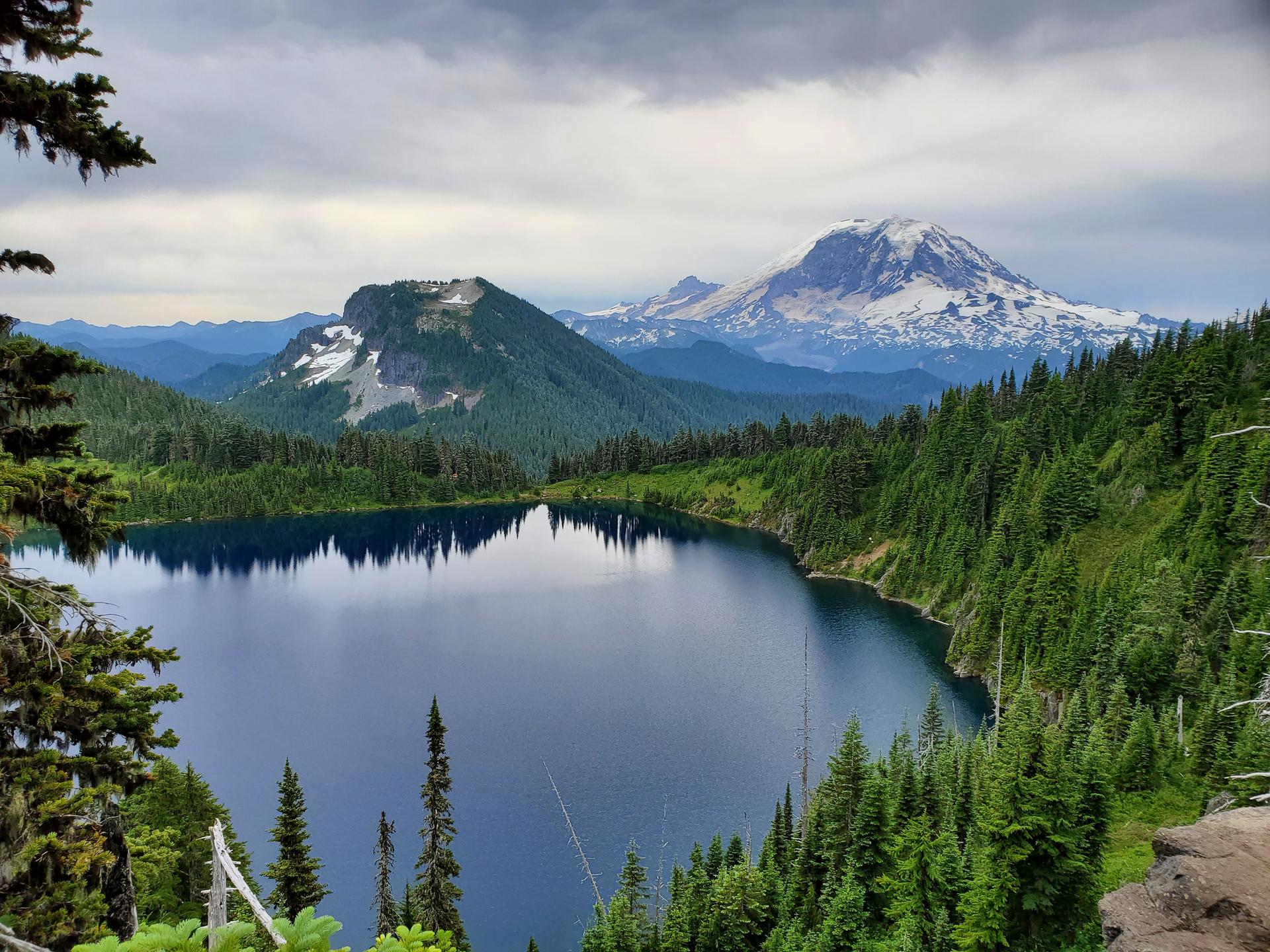 Panoramic view of a large blue lake surrounded by forests and snow-capped mountains.