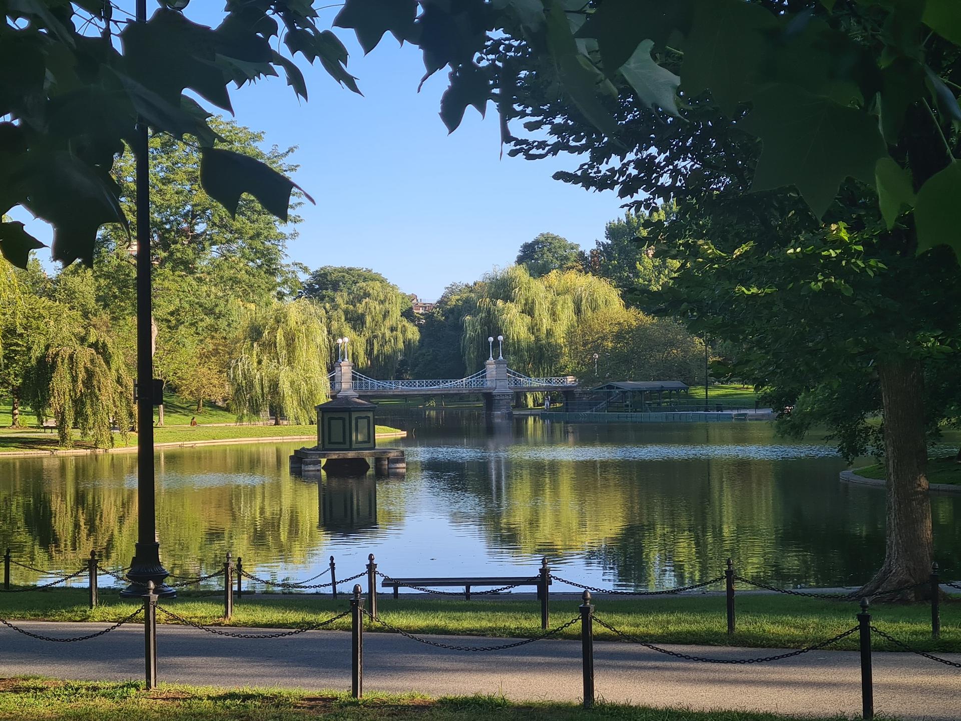 Pond with a decorative bridge and boat house, framed by green trees, sunny.