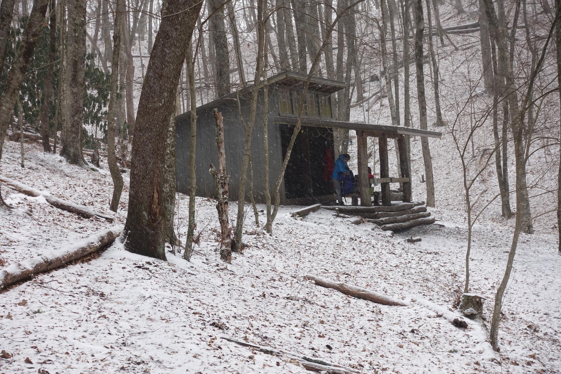 Rustic wooden shelter in a snowy forest with two people inside.