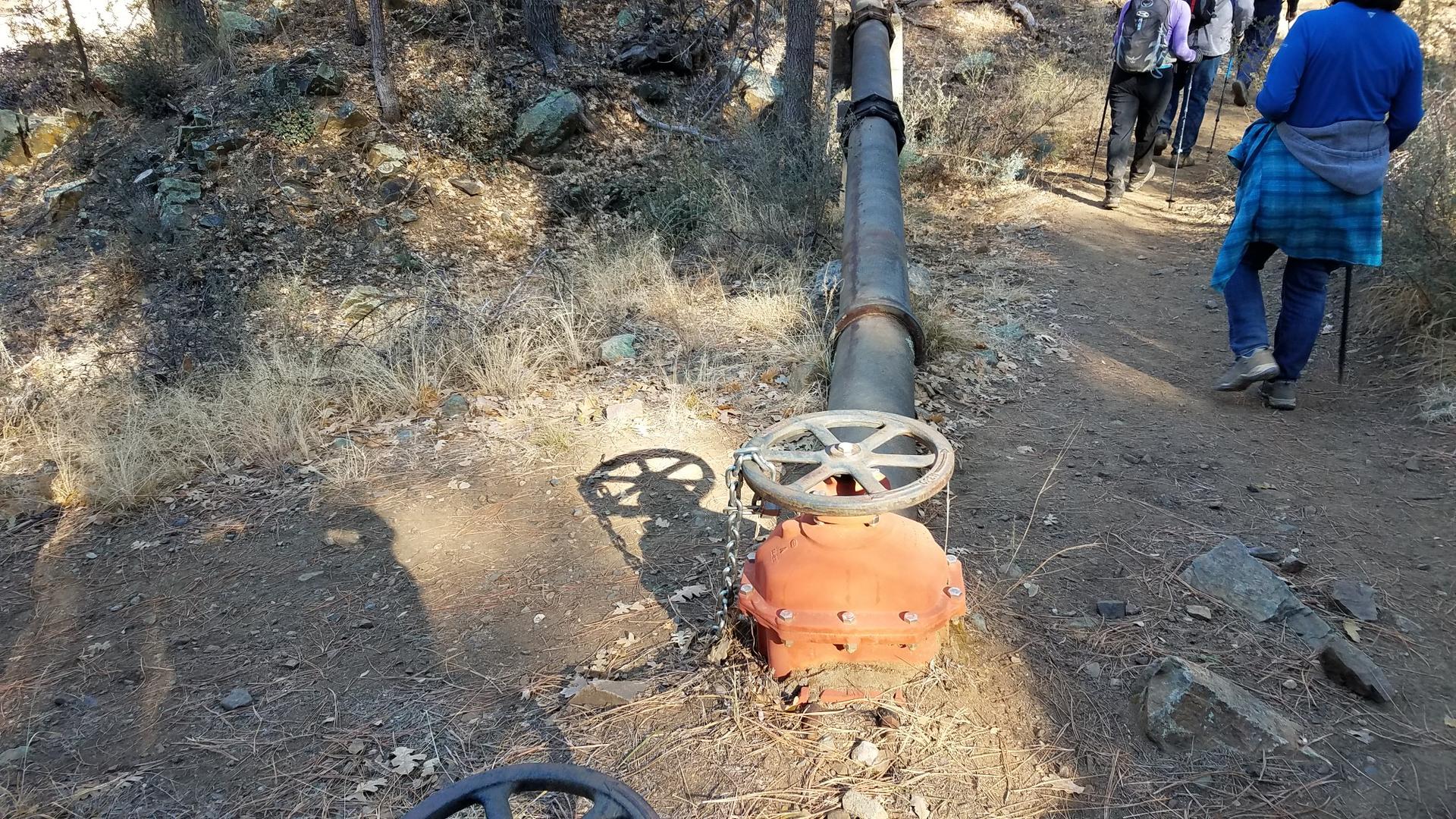 Hikers walk past a large metal pipe with a valve on a dirt trail.