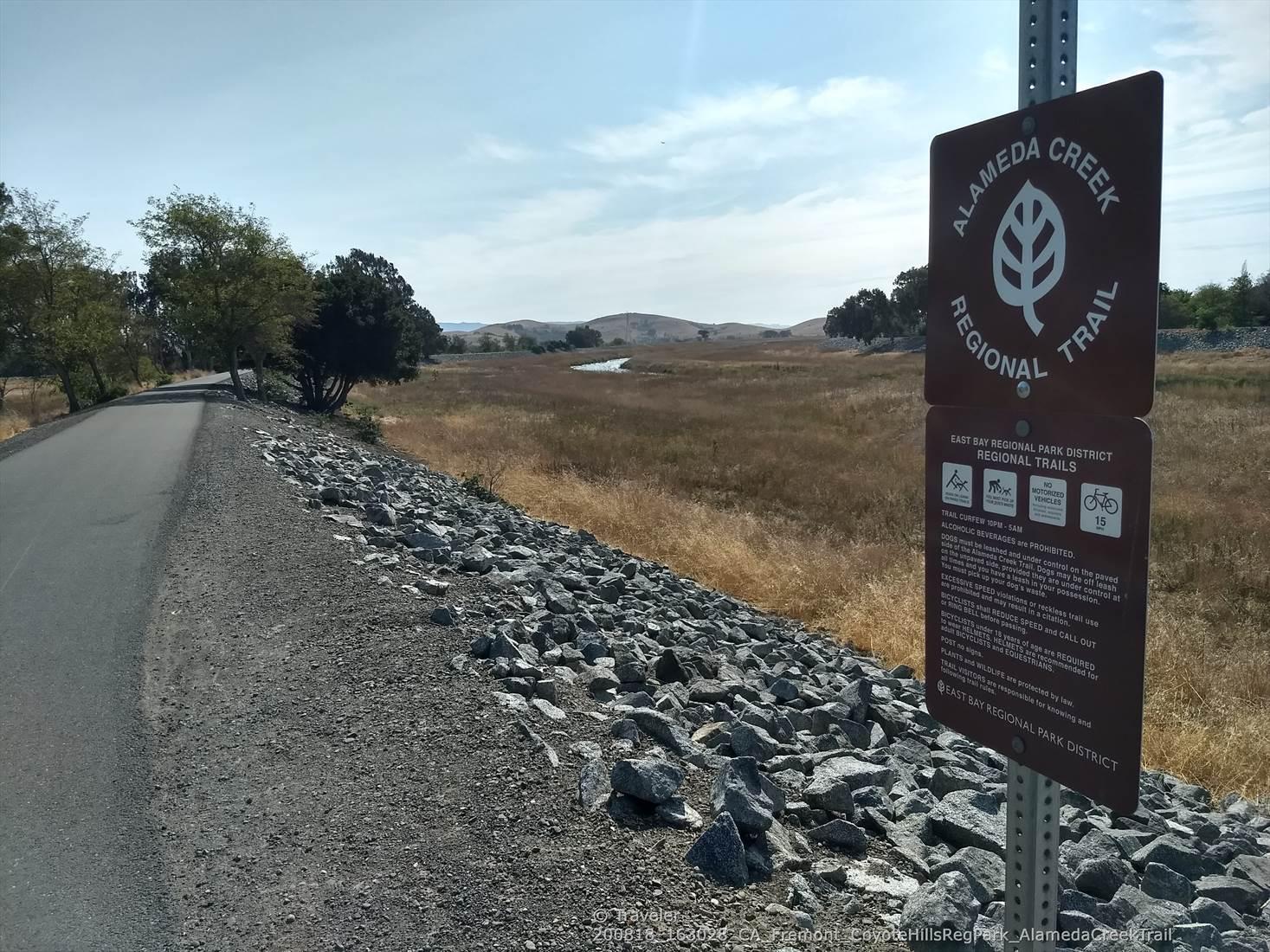 Trail sign beside a paved path, with dry fields and distant hills.